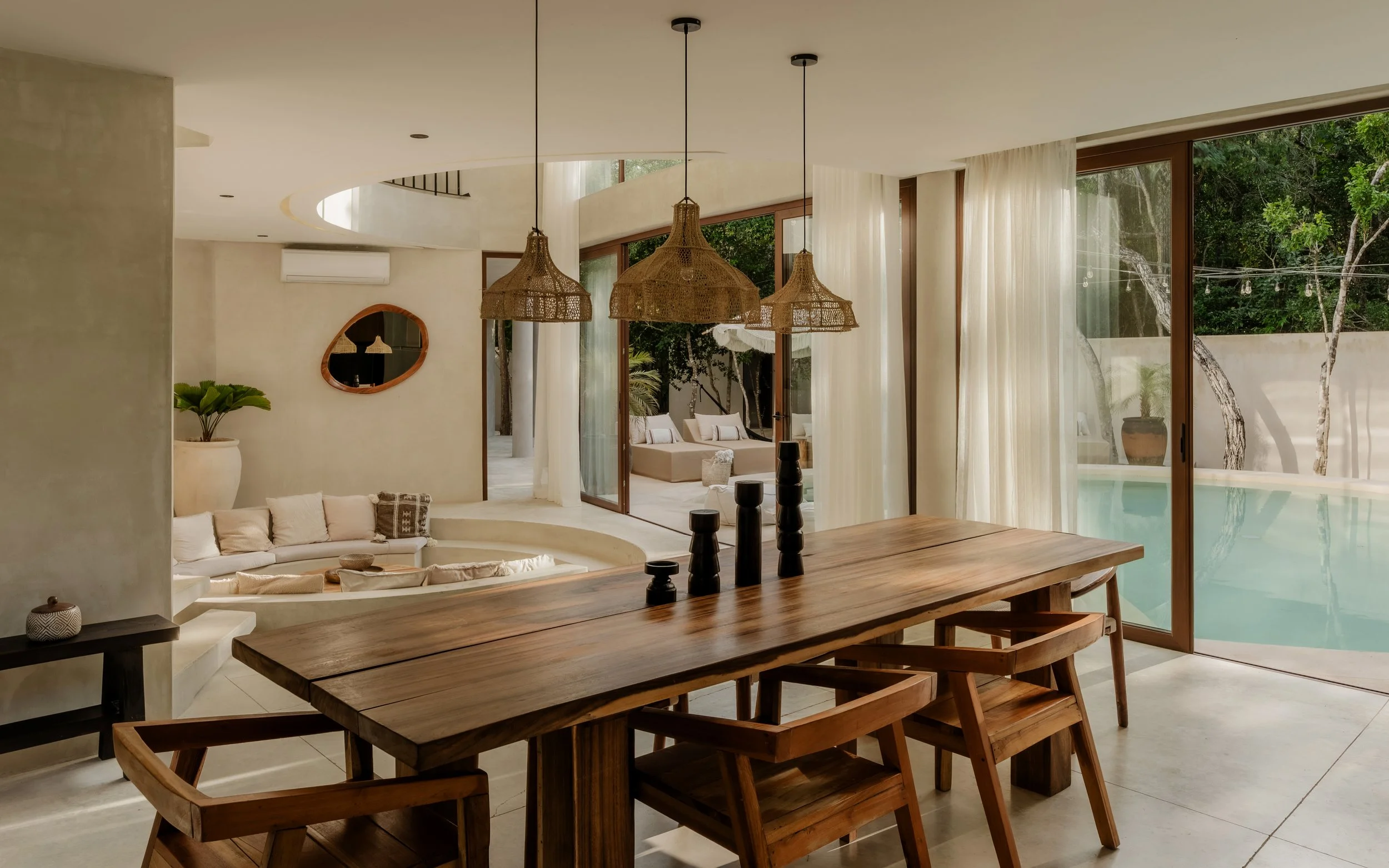 A modern open-concept living space with a wooden dining table, woven pendant lights, a curved built-in sofa, potted plant, large floor-to-ceiling windows with curtains, and a view of trees and a pool outside.