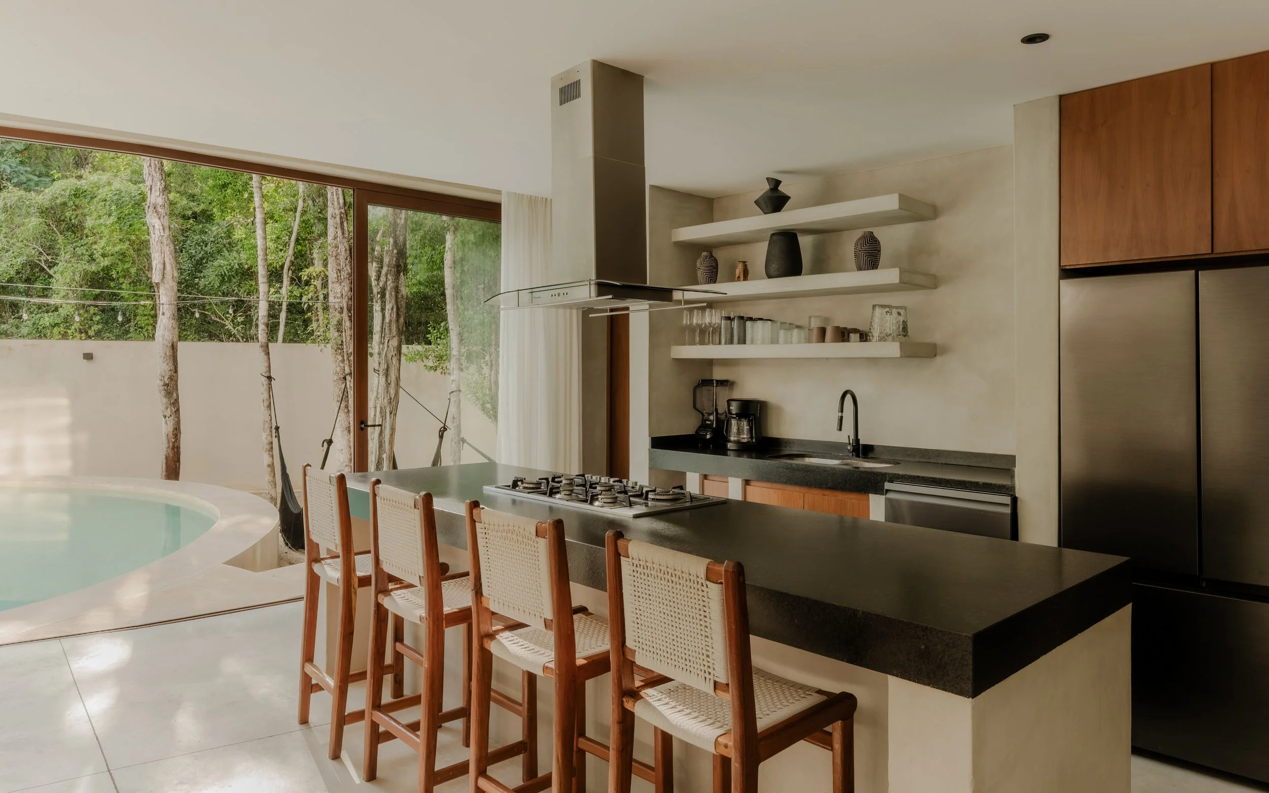 Modern kitchen with a large window overlooking a wooded outdoor area, featuring a kitchen island with chairs, open shelves with decorated vases, a countertop with a coffee maker, and a stainless steel refrigerator.