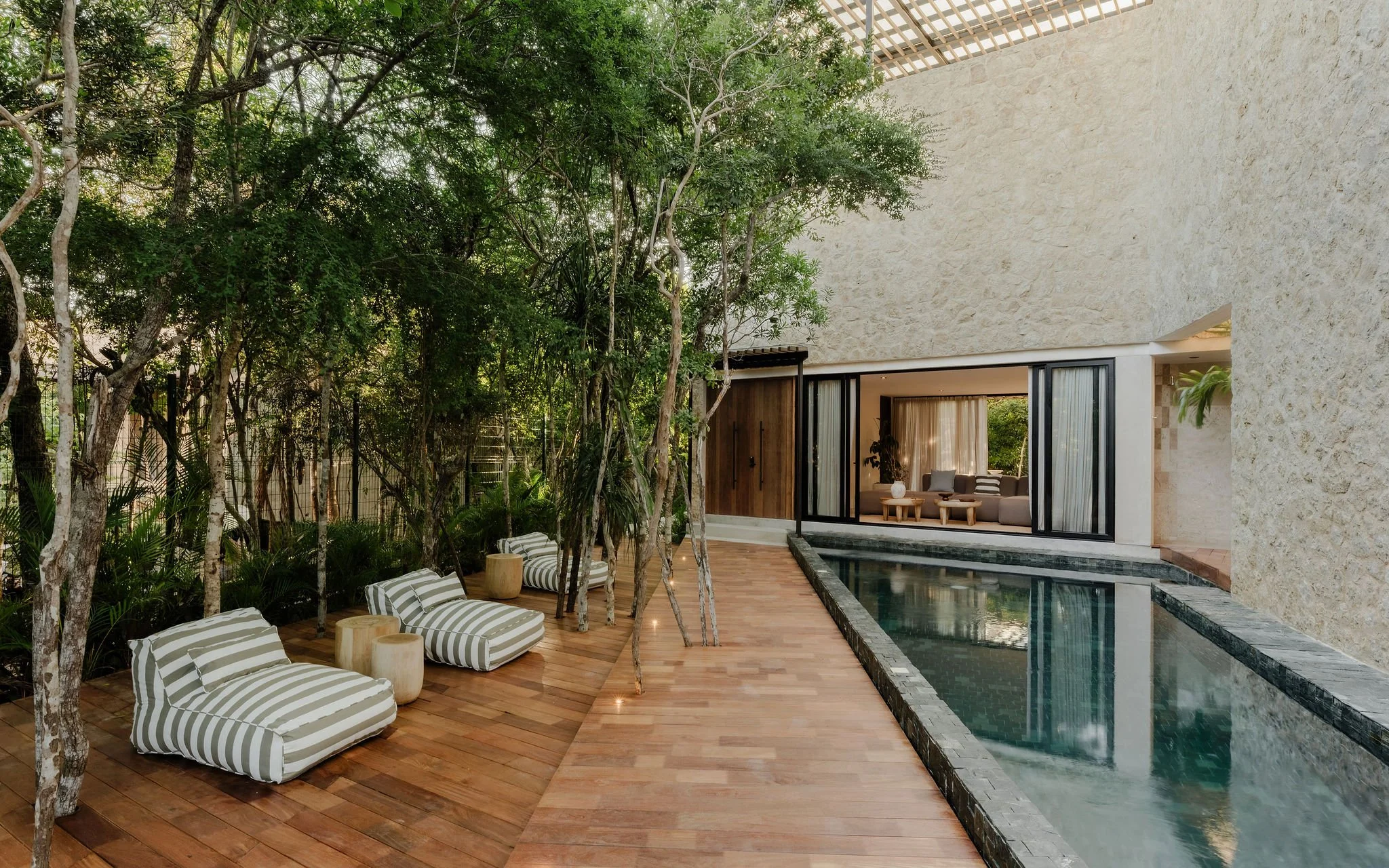Modern house with open living area, large windows, and a swimming pool, surrounded by lush green trees.