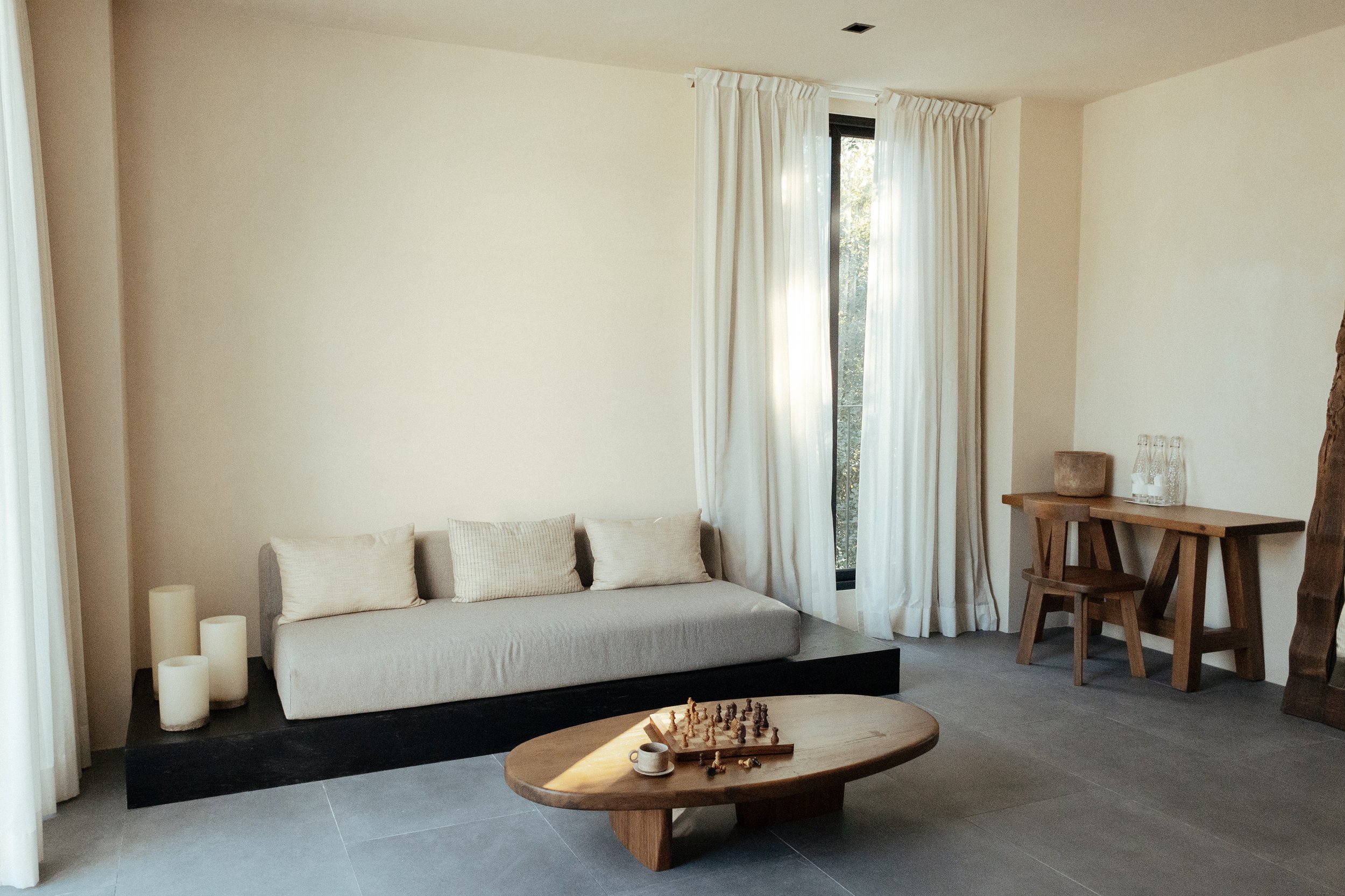 A modern living room with a gray sofa, white cushions, and a black rug. There are white candles on the floor, a wooden coffee table with a chess set and coffee cups, and a window with white curtains. A wooden table with glass bottles and a ceramic pot is against the wall.