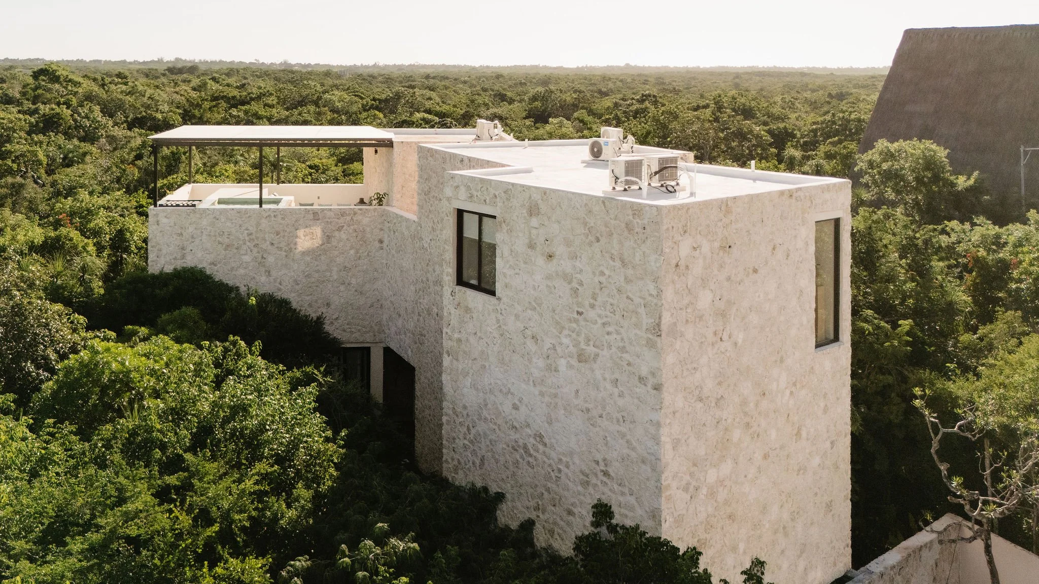 A modern white stone building surrounded by lush green trees, with air conditioning units on the roof and a small balcony with a hot tub.