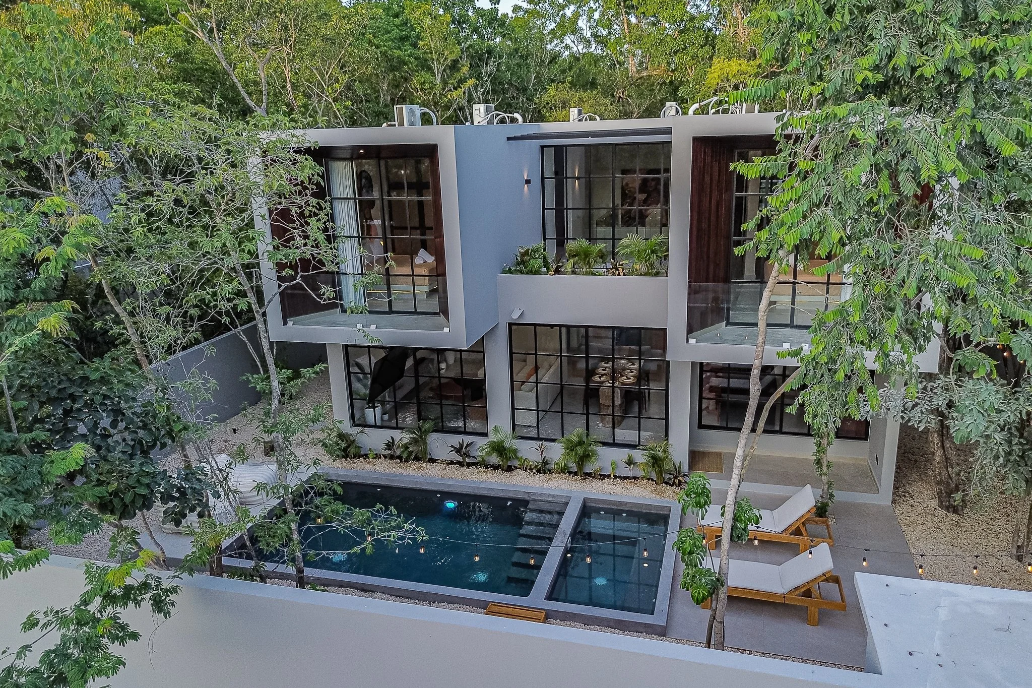 Modern multi-story house with large glass windows, a swimming pool, outdoor seating, and surrounded by green trees.