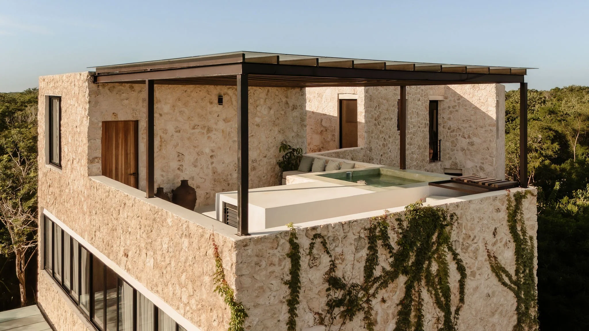 A modern stone house with an outdoor balcony featuring a small pool, seating area, and a pergola on top, surrounded by trees.