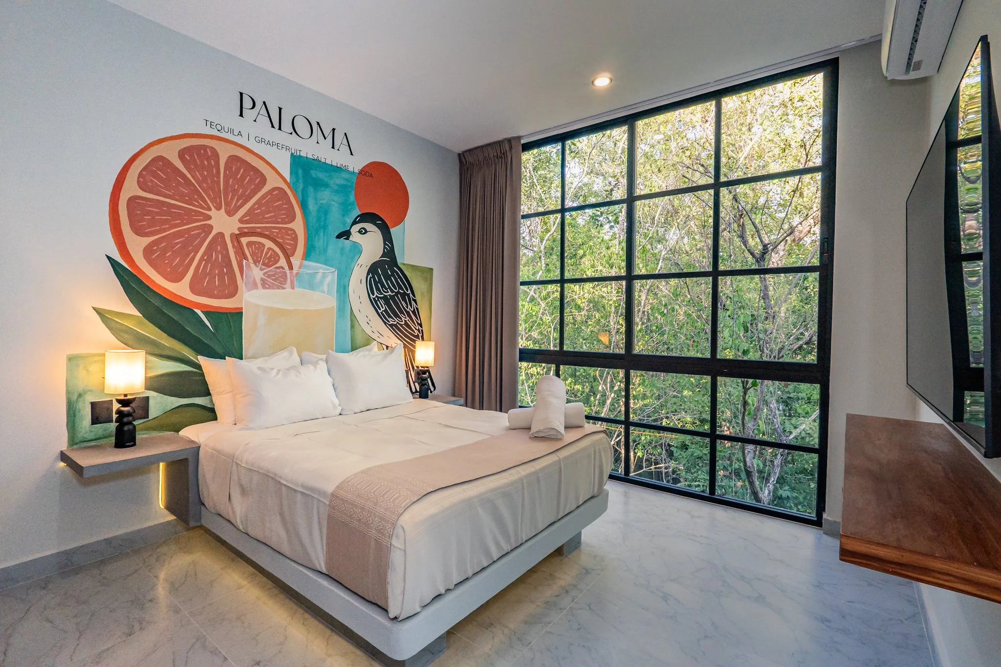 A modern bedroom with a large window showing trees outside, a bed with white linens, a painted wall art featuring a grapefruit and a bird, bedside lamps, and a TV mounted on the wall.