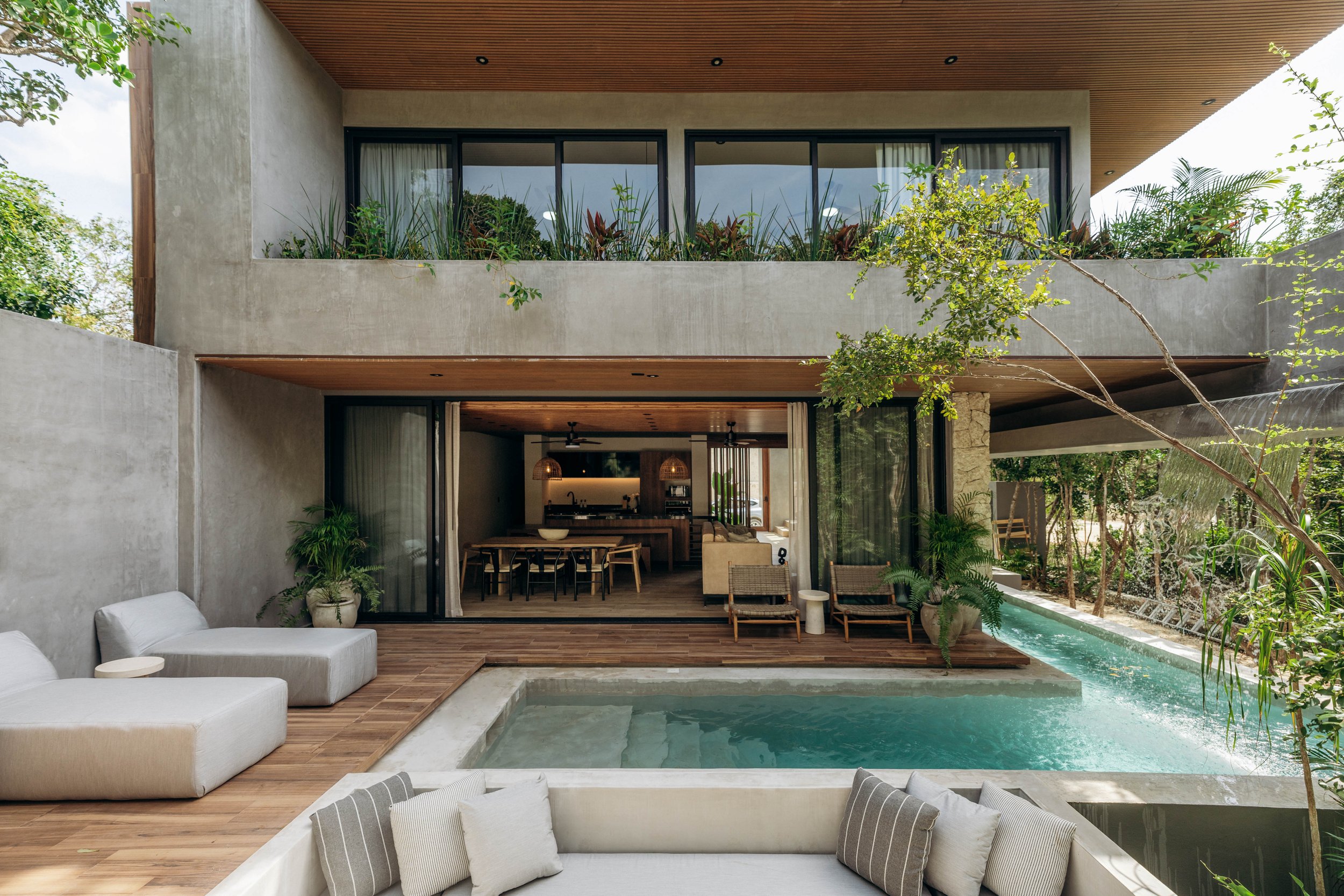 Modern house with large balcony filled with plants and glass doors leading inside. There is a swimming pool in the foreground surrounded by wooden decking, with outdoor furniture including white cushioned seating and pillows.