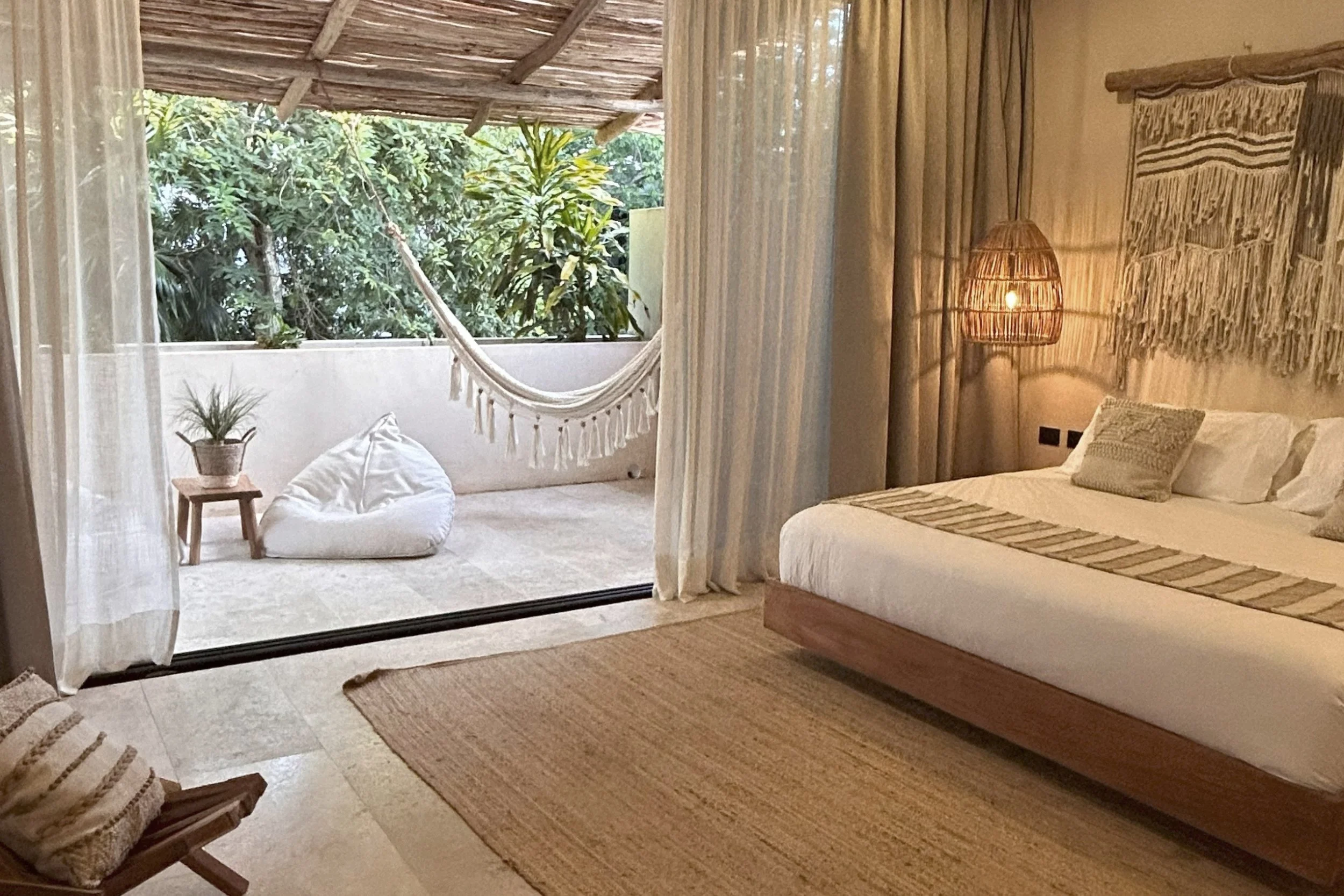 A bedroom with an outdoor balcony. The bed has a beige bedspread and pillows. To the right is a woven wall hanging and a rattan floor lamp. The balcony has a hammock, small side table with a potted plant, and lush green trees outside.