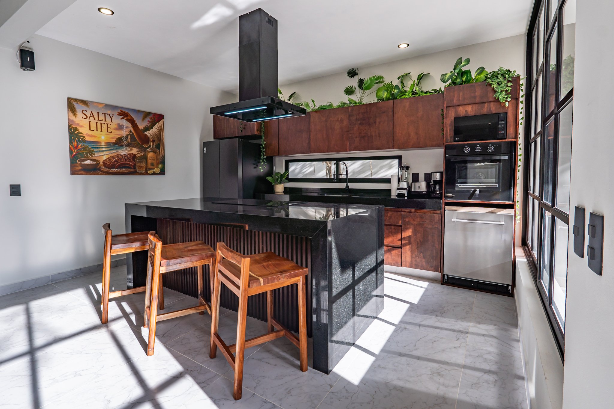 Modern kitchen with wooden cabinets, black countertops, and plants on top of cabinets. There are wooden chairs at a black island, a wall-mounted oven, microwave, blender, and a framed poster that reads 'Salty Life' with a sunset scene.