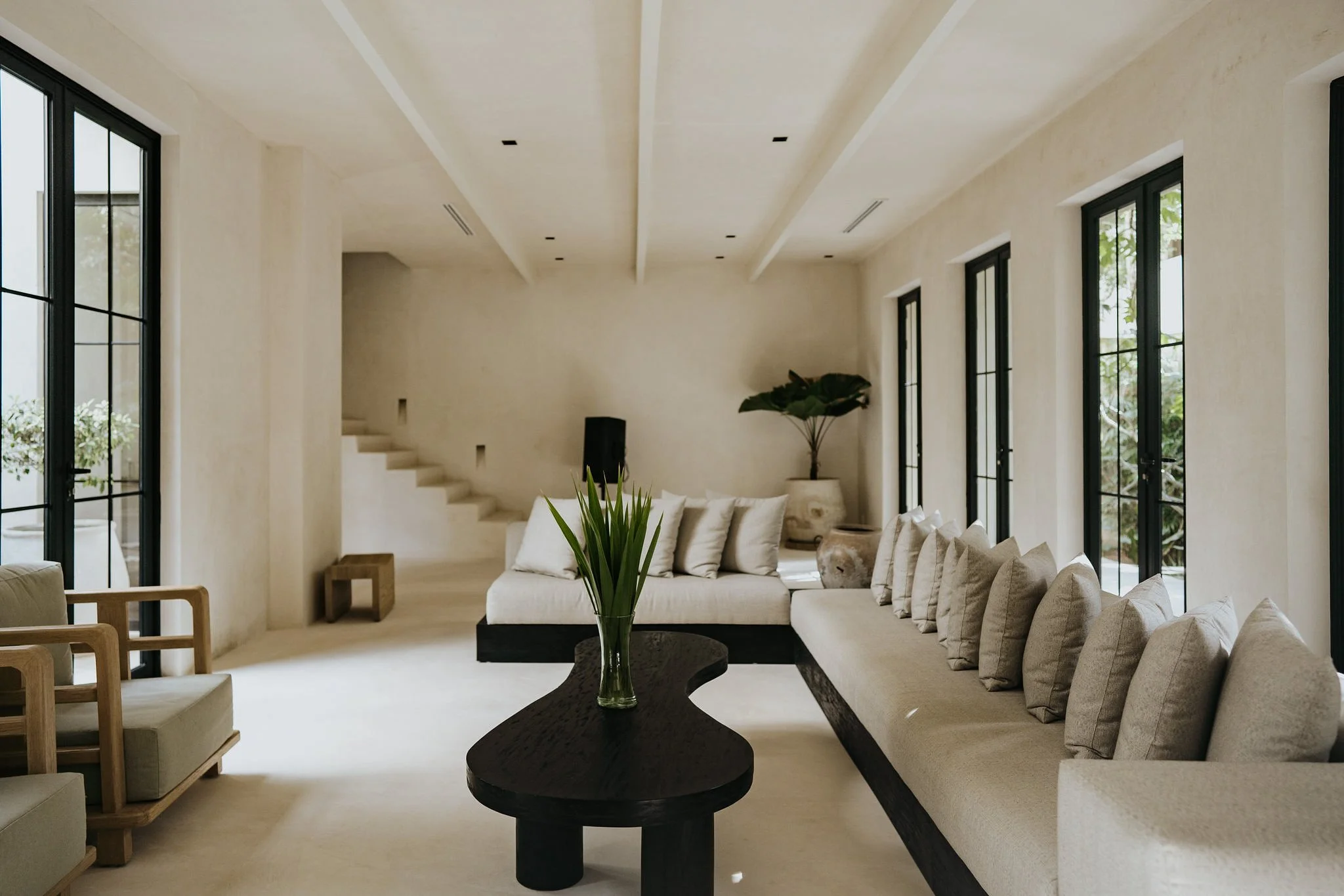 Modern living room with beige walls and ceiling, large windows with black frames, white and beige sofas, a black coffee table with a green plant in a glass vase, and decorative items.