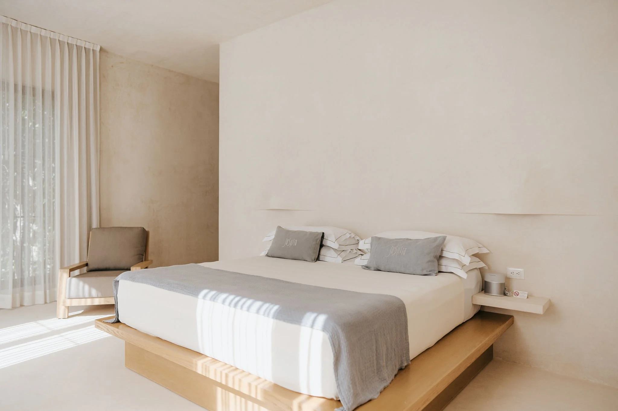 A modern bedroom with a minimalistic design featuring a white bed with gray and white pillows, a small bedside table with a speaker, and a beige armchair next to a window with white curtains.