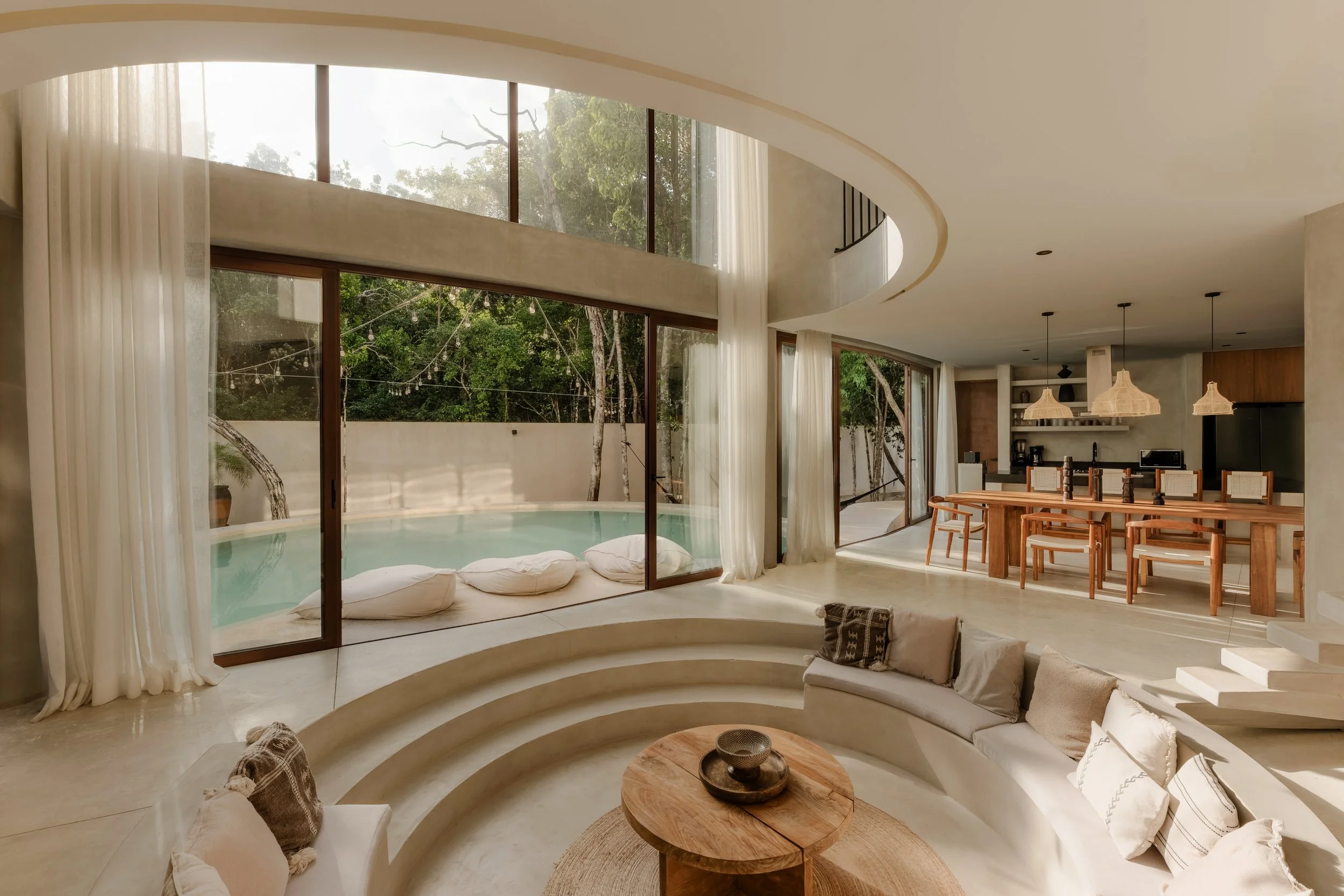 Modern living room with large curved windows overlooking trees, white curtains, round seating with cushions, a wooden coffee table, and a dining area with hanging wicker lights.