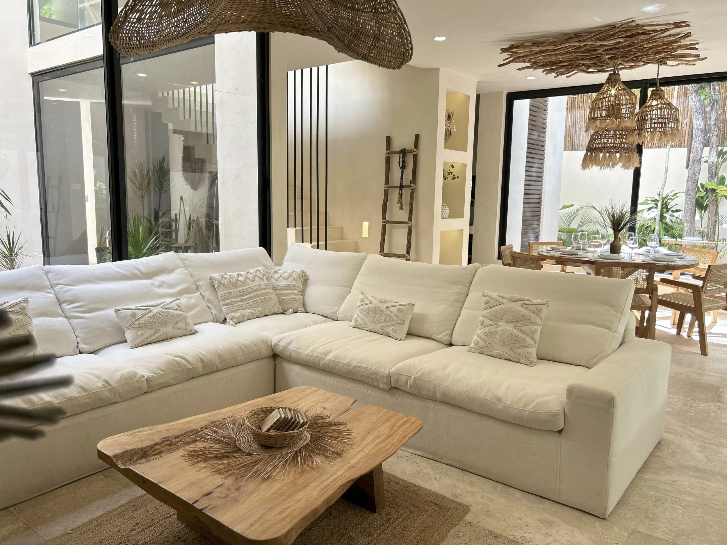 Living room with white sectional sofa, wooden coffee table, and dining area with wooden table and woven chairs, decorated with bamboo and wicker pendant lights and large windows showing outdoor greenery.