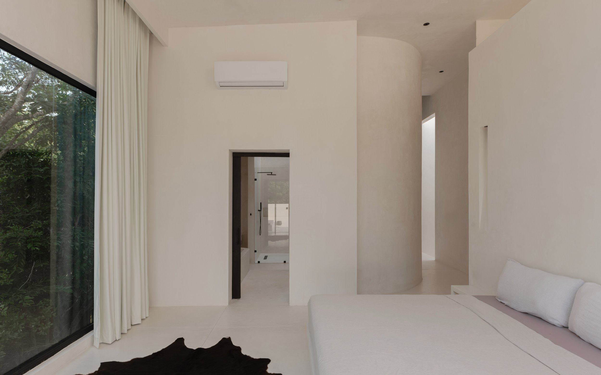 Minimalist interior with white walls, large glass sliding door with white curtains, a cowhide rug, and a white cushioned seating area with pillows.