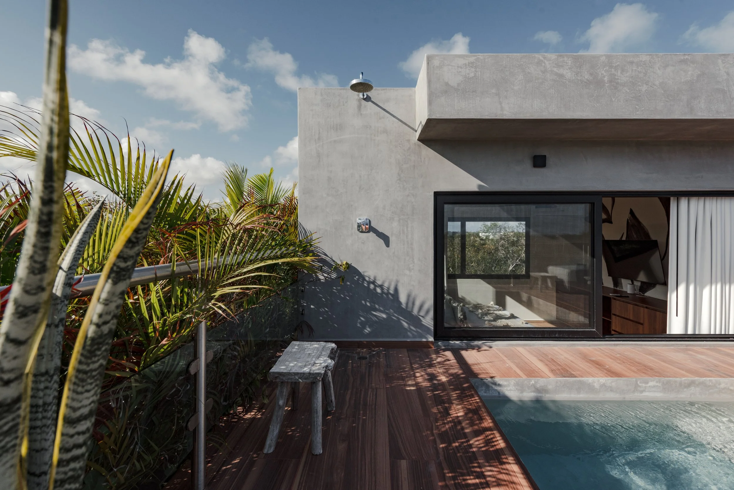 Modern house with gray exterior wall, large glass sliding door, outdoor showerhead, and view of a swimming pool on a wooden deck, with lush greenery and a blue sky with clouds.