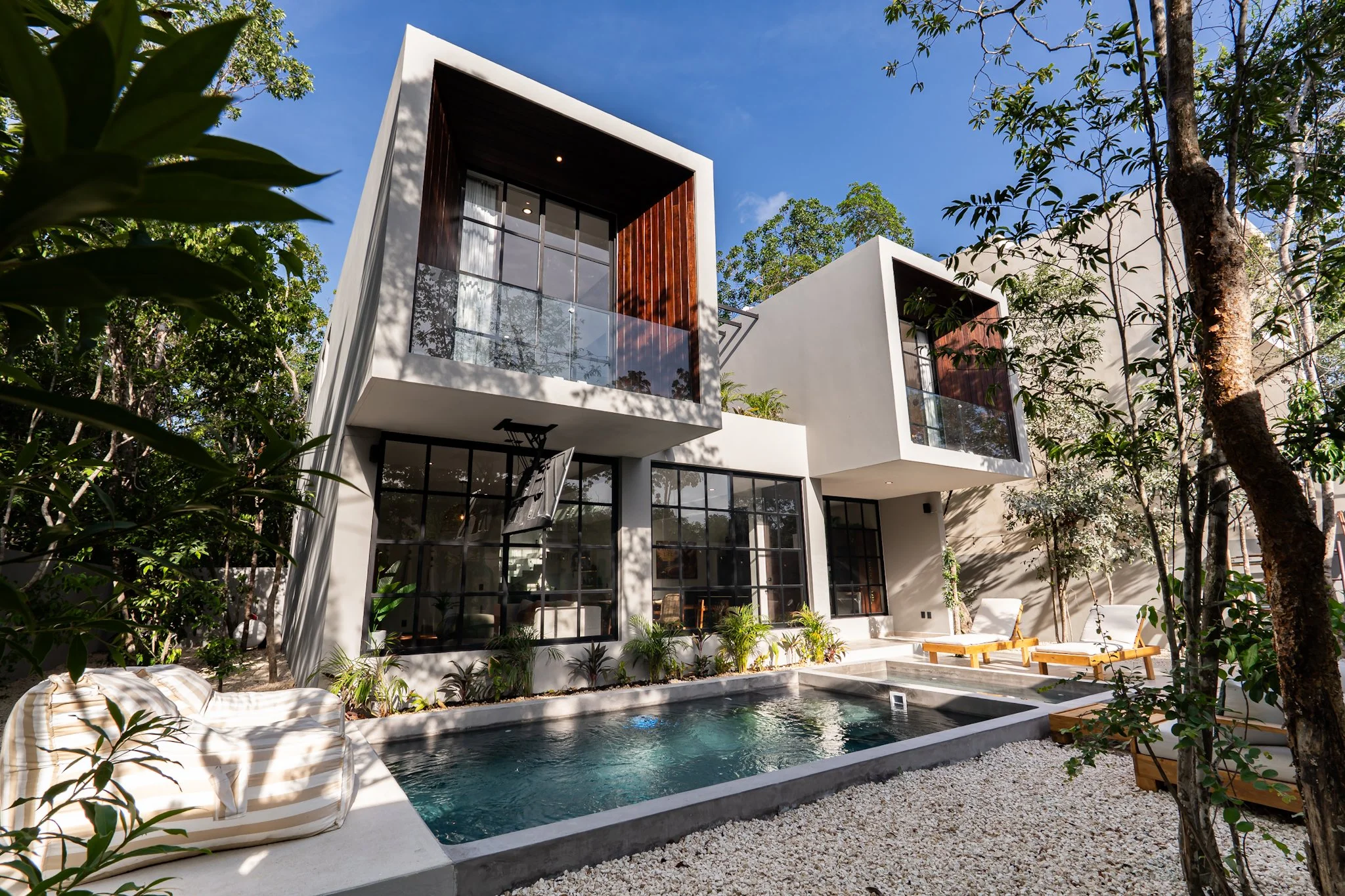 Modern white residential building with large glass windows, a balcony, and wooden accents, surrounded by trees, with a swimming pool and lounge chairs in the yard.