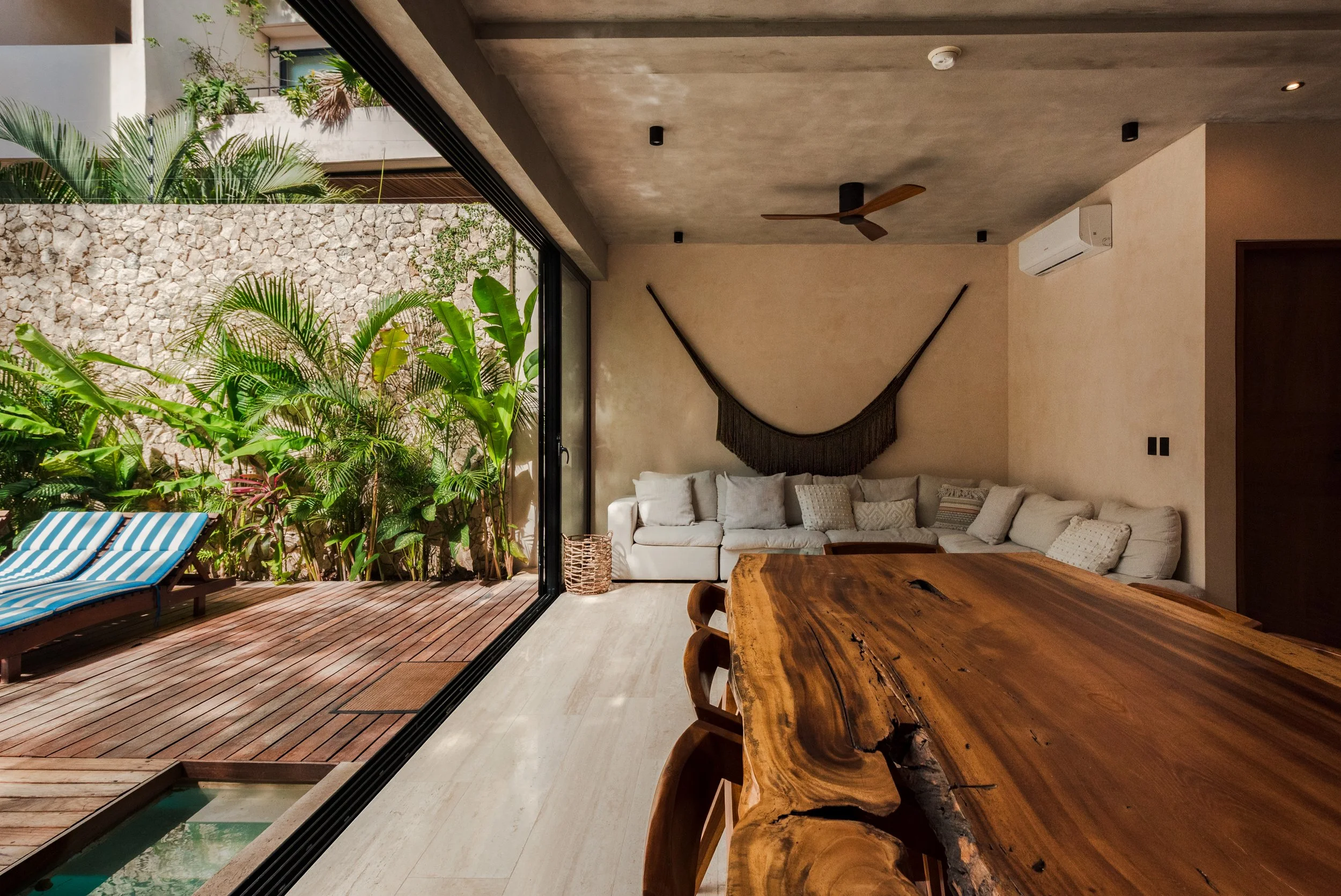 Indoor living space opening to a wooden deck with lush tropical plants, a stone wall, and the outdoors. Inside features a white sectional sofa, a wooden dining table, modern ceiling fan, air conditioning unit, and a beige wall with black wall-mounted lights.