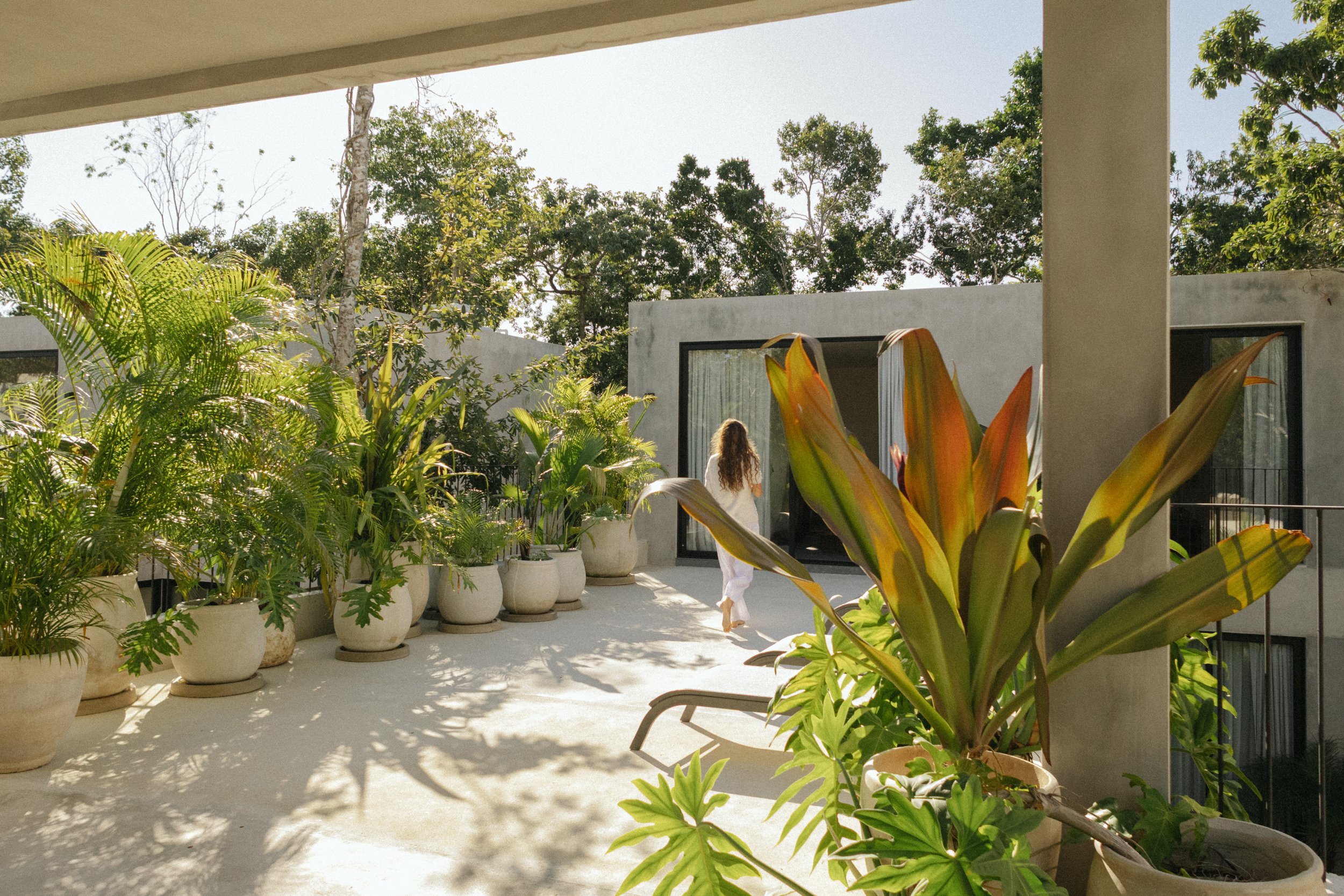 A person with long curly hair wearing white pants and a white top walking on a patio surrounded by potted plants, with a backdrop of modern houses and tall trees.