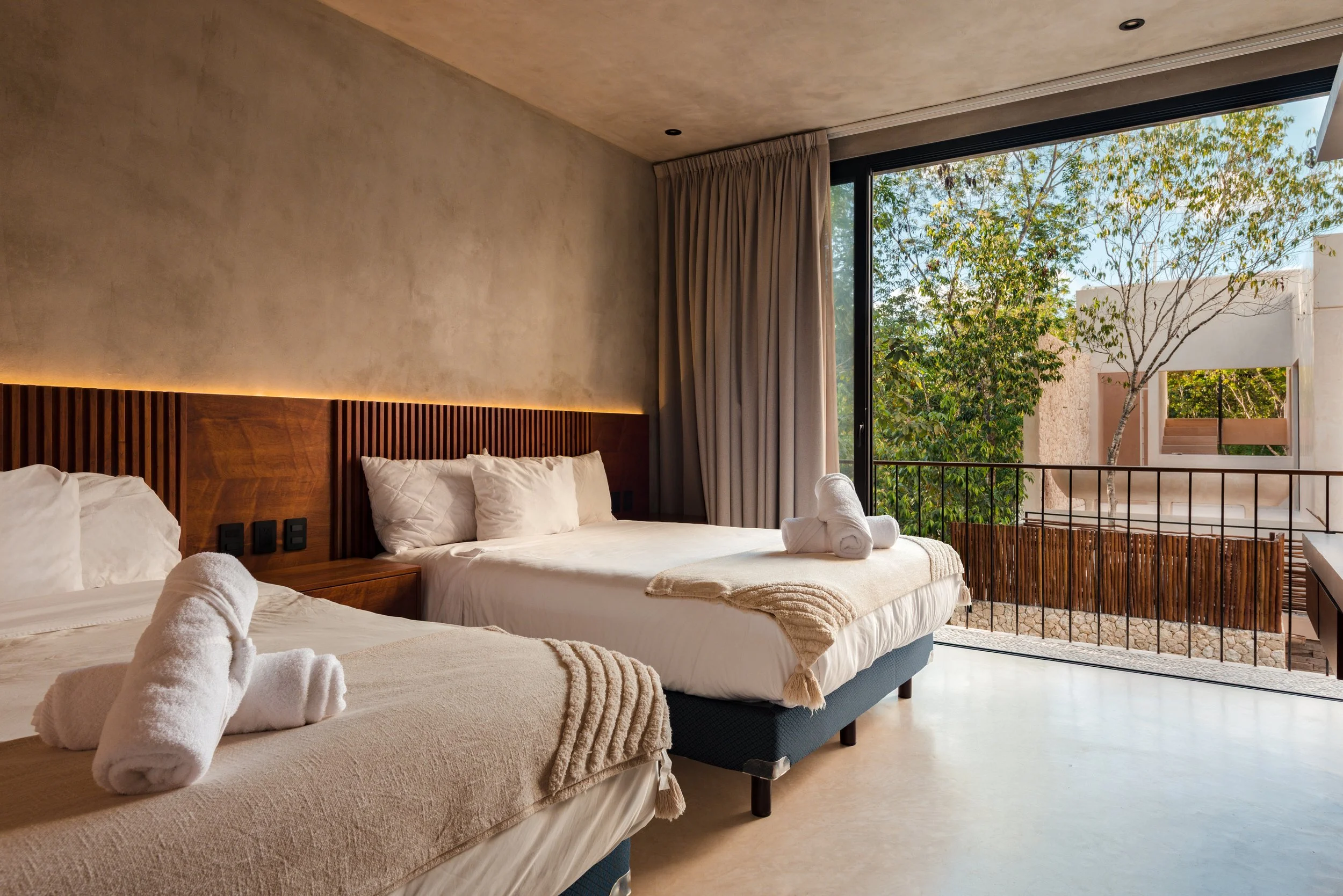Bedroom with two beds, white towels folded on each, beige walls, large window with curtains, view of outside trees and modern building.