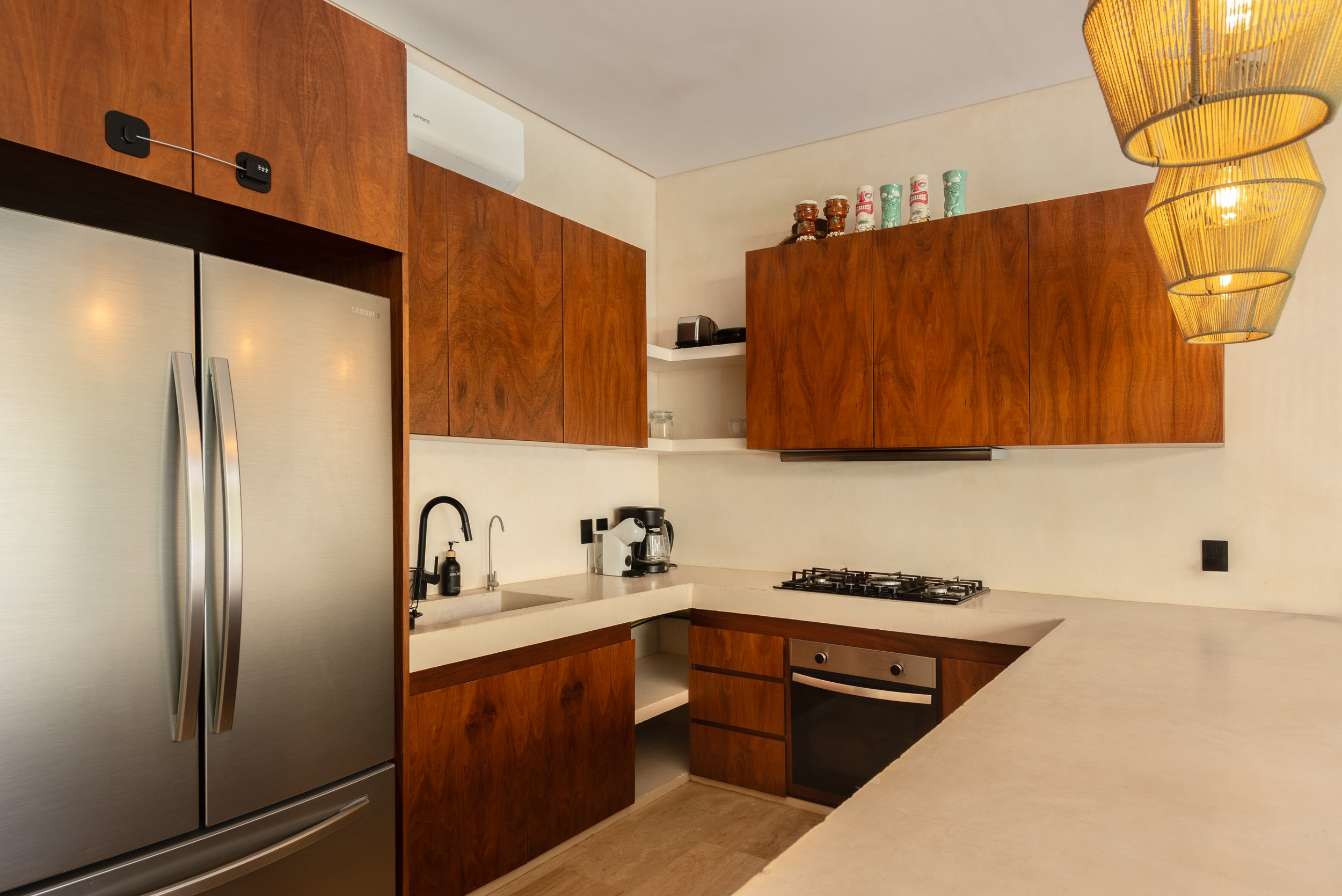 Modern kitchen with wooden cabinets, a stainless steel refrigerator, a coffee maker, and a gas stove. Yellow pendant lights hanging from the ceiling, and a white countertop with a sink and black faucet.