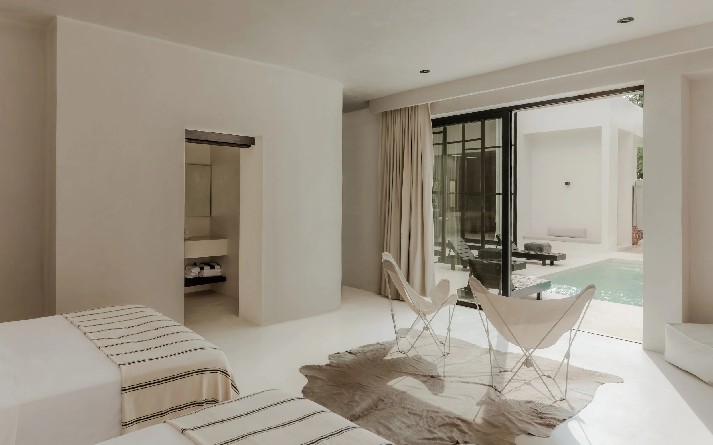 Bright modern living room with large glass doors opening to a pool area, white walls, beige curtains, and minimal furniture including white chairs and a cowhide rug.