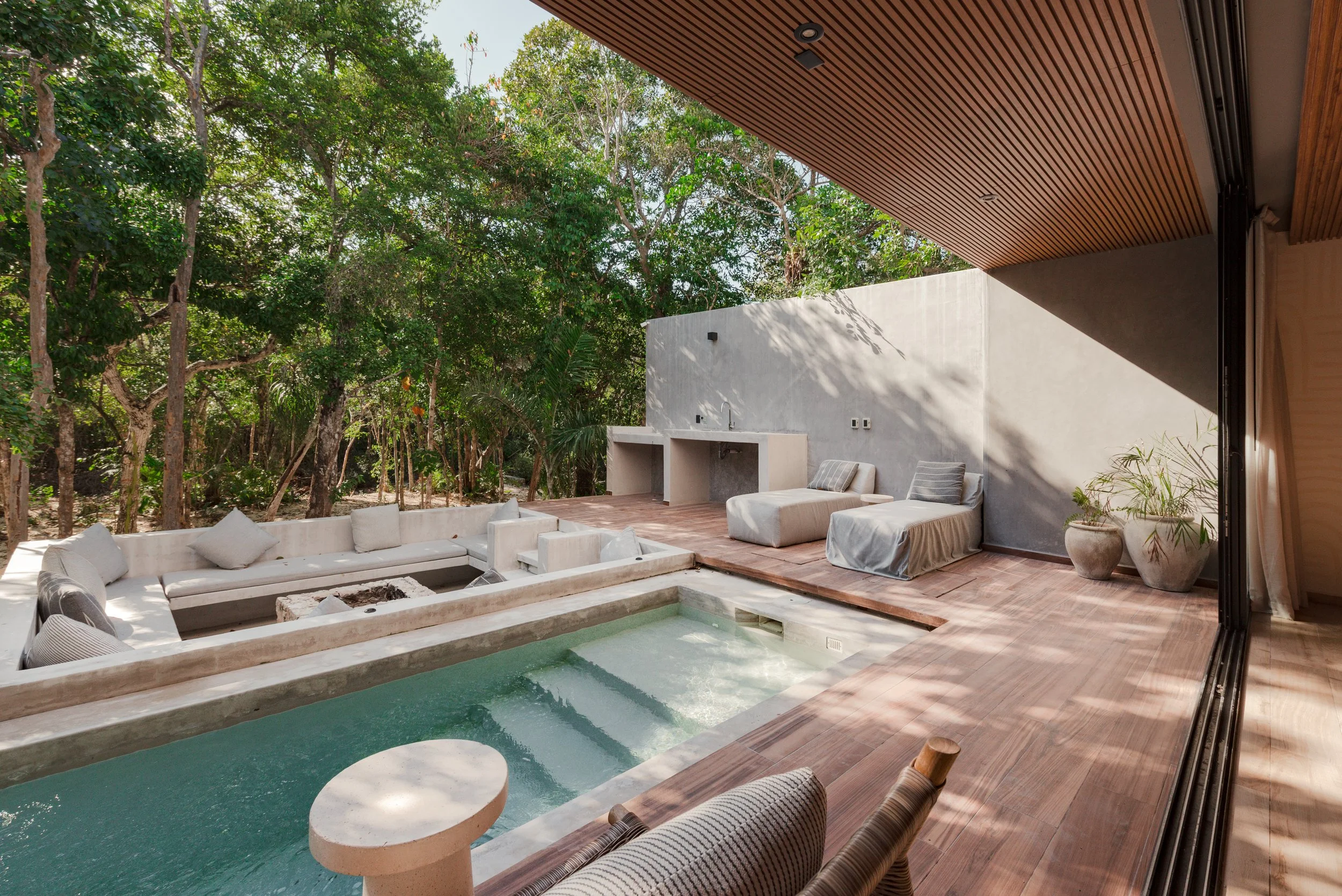 Modern outdoor patio with white cushioned seating area, small pool, and a wall with a built-in outdoor sink. Surrounded by lush green trees and shaded by a wooden ceiling.