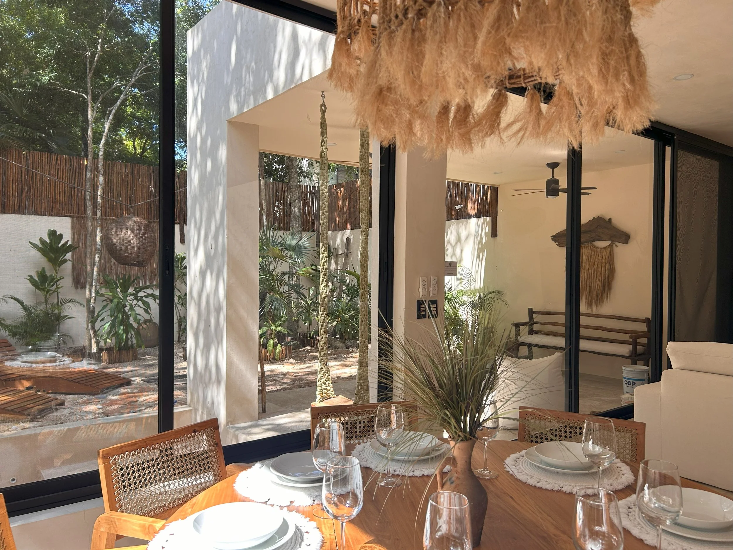 Interior of a modern home with large glass windows, view of a lush garden, wooden dining table set with glassware and white plates, and decorative hanging plants.