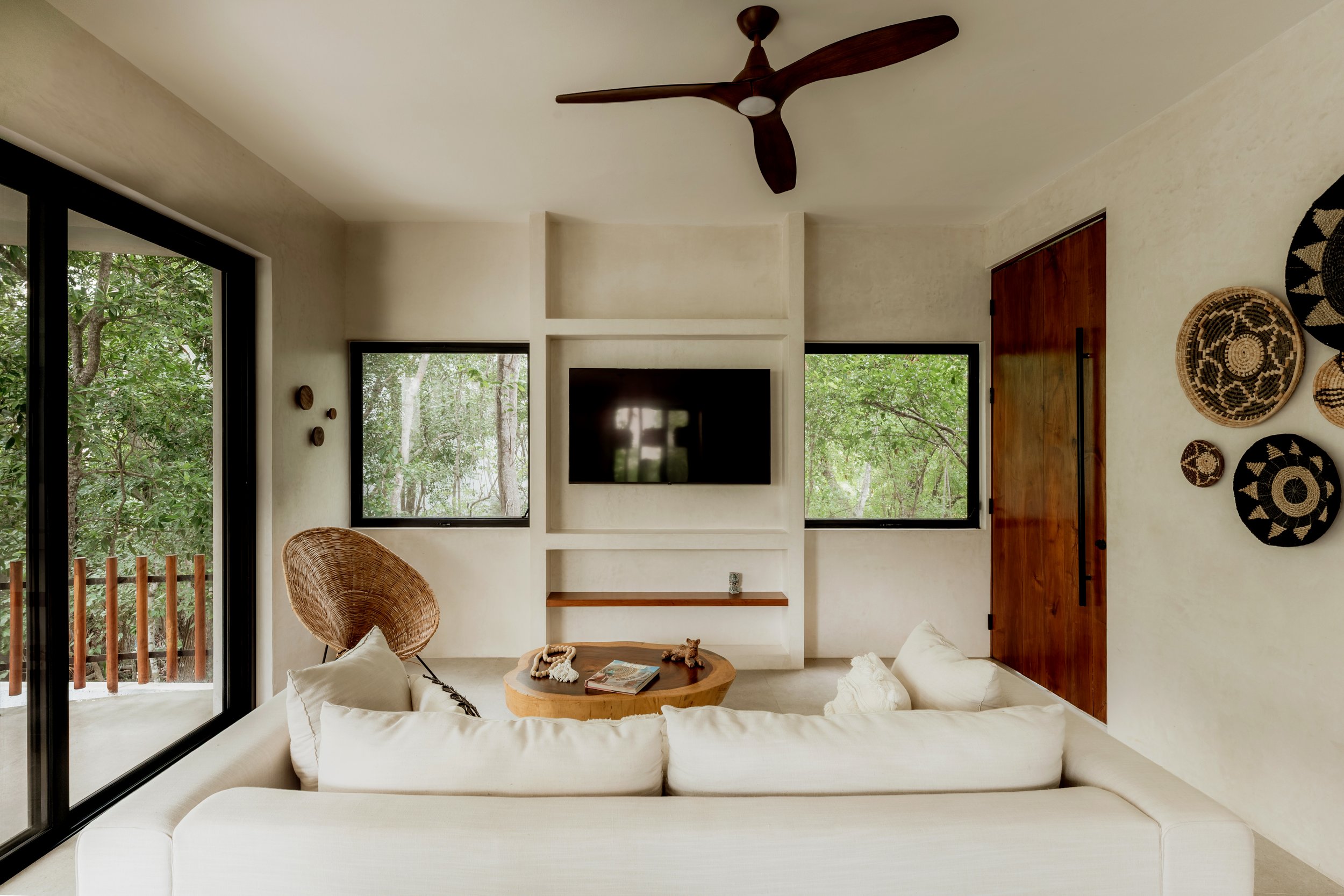 Living room with large windows showing trees outside, white couches, a round wooden coffee table with books, and a woven chair, with wall decor and a ceiling fan.