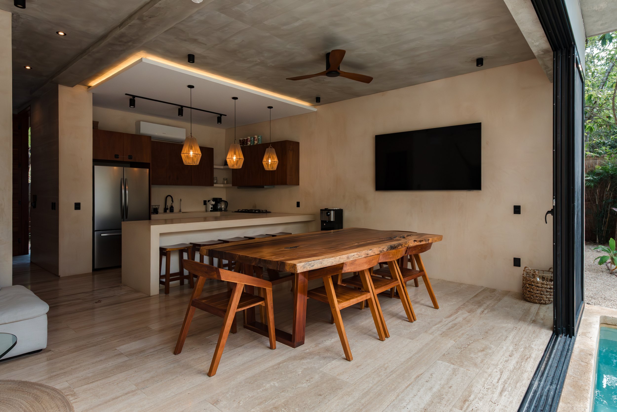 Open-plan kitchen and dining area with a large wooden table, ultrasound chairs, pendant lights, kitchen cabinets, stainless steel refrigerator, TV, ceiling fan, and sliding glass doors opening to outdoor greenery.