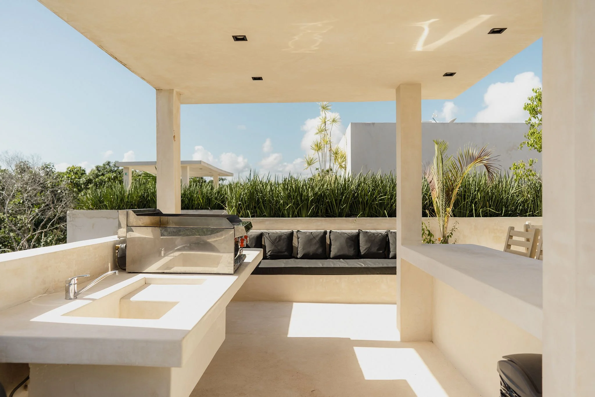 An outdoor patio with a built-in sink, a grill, black cushioned seating, and a view of trees and blue sky.