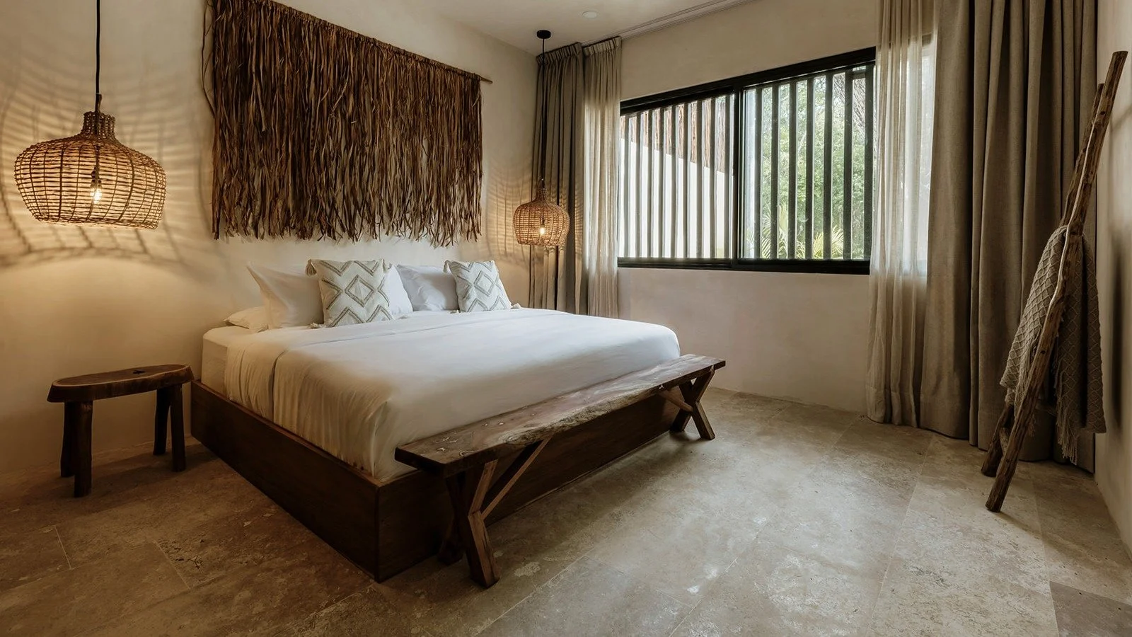 A bedroom with a large bed, white bedding, and pillows, flanked by rattan pendant lamps. The room has a large window with blinds and curtains, and a rustic wooden bench at the foot of the bed.