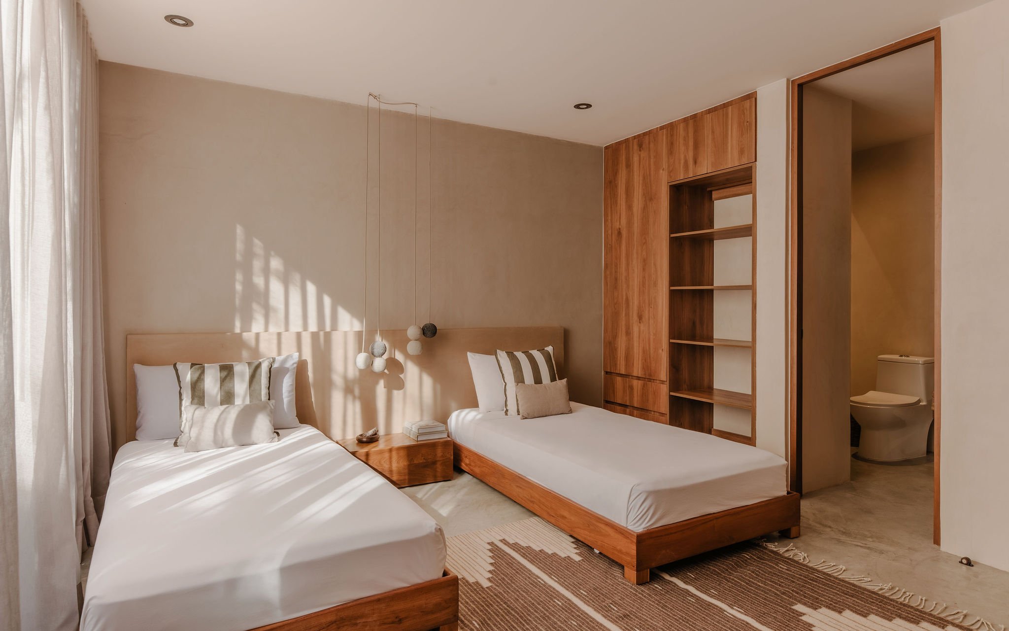 A modern bedroom with two beds, white bedding, striped pillows, a hanging pendant light with multiple globes, a wooden nightstand, built-in wooden shelves, and a partial view of a bathroom with a toilet.