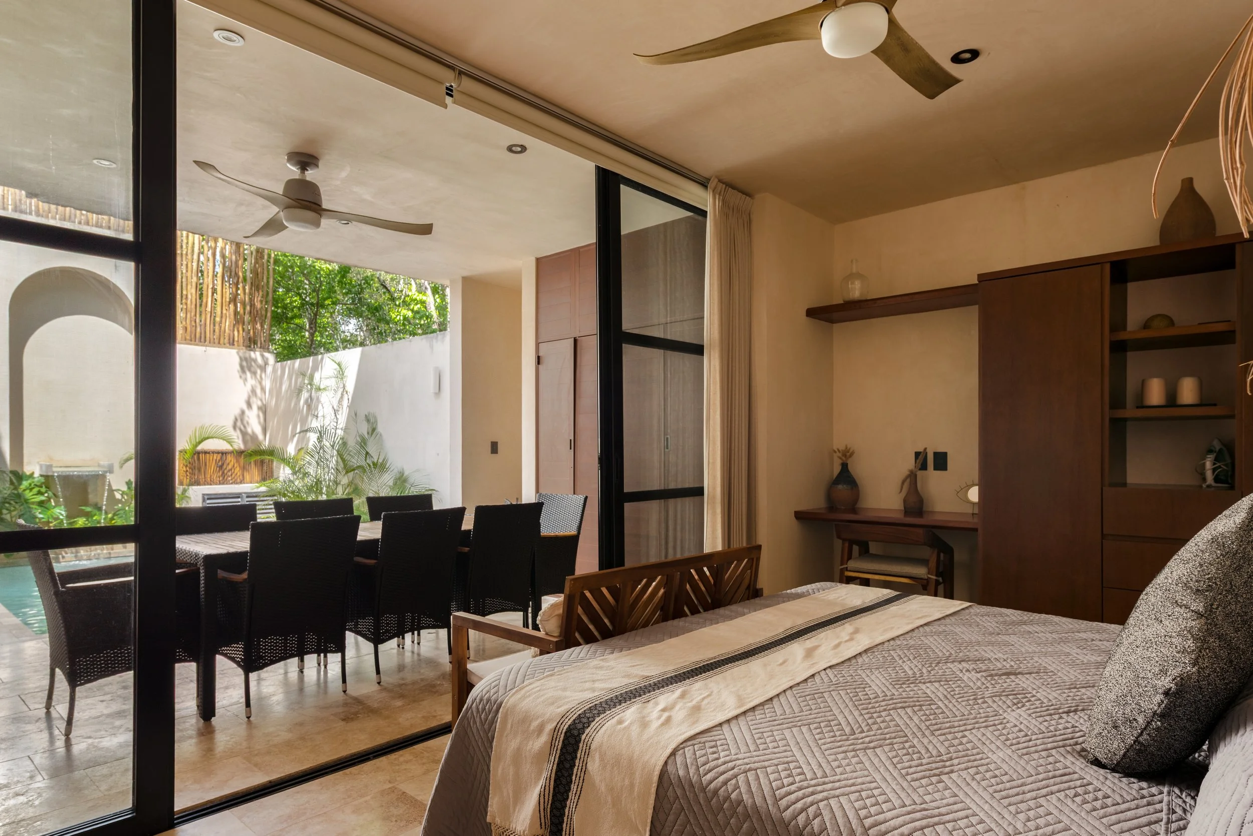 View from inside a bedroom looking out through sliding glass doors to an outdoor dining area with a black table and six chairs. The outdoor space features greenery and a bamboo fence. Inside, there is a wooden bed with a beige and black quilt, a wooden shelf with vases and candles, and a ceiling fan.