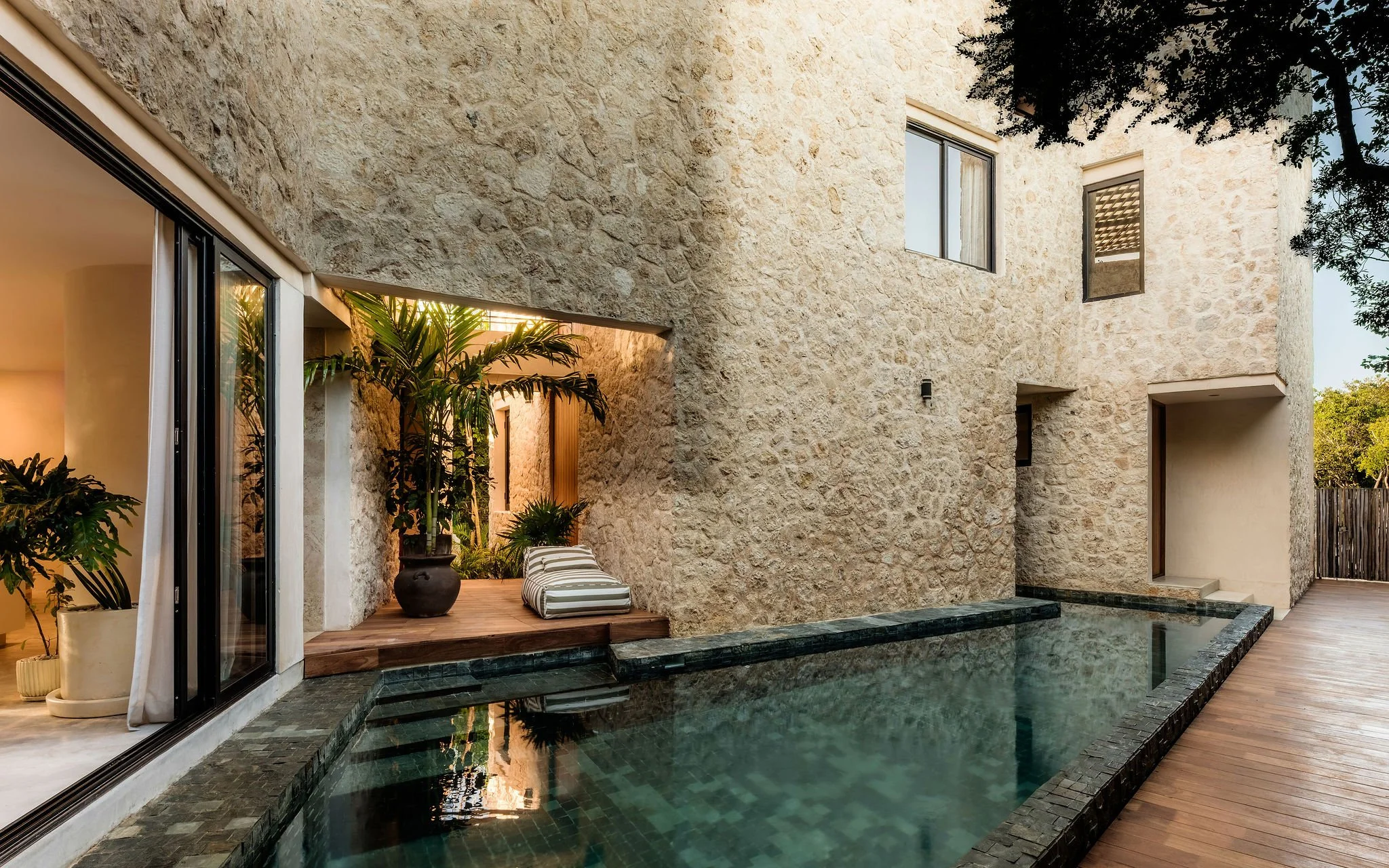 A modern house with a textured stone exterior wall, large windows, and a patio area with plants, outdoor furniture, and a swimming pool.