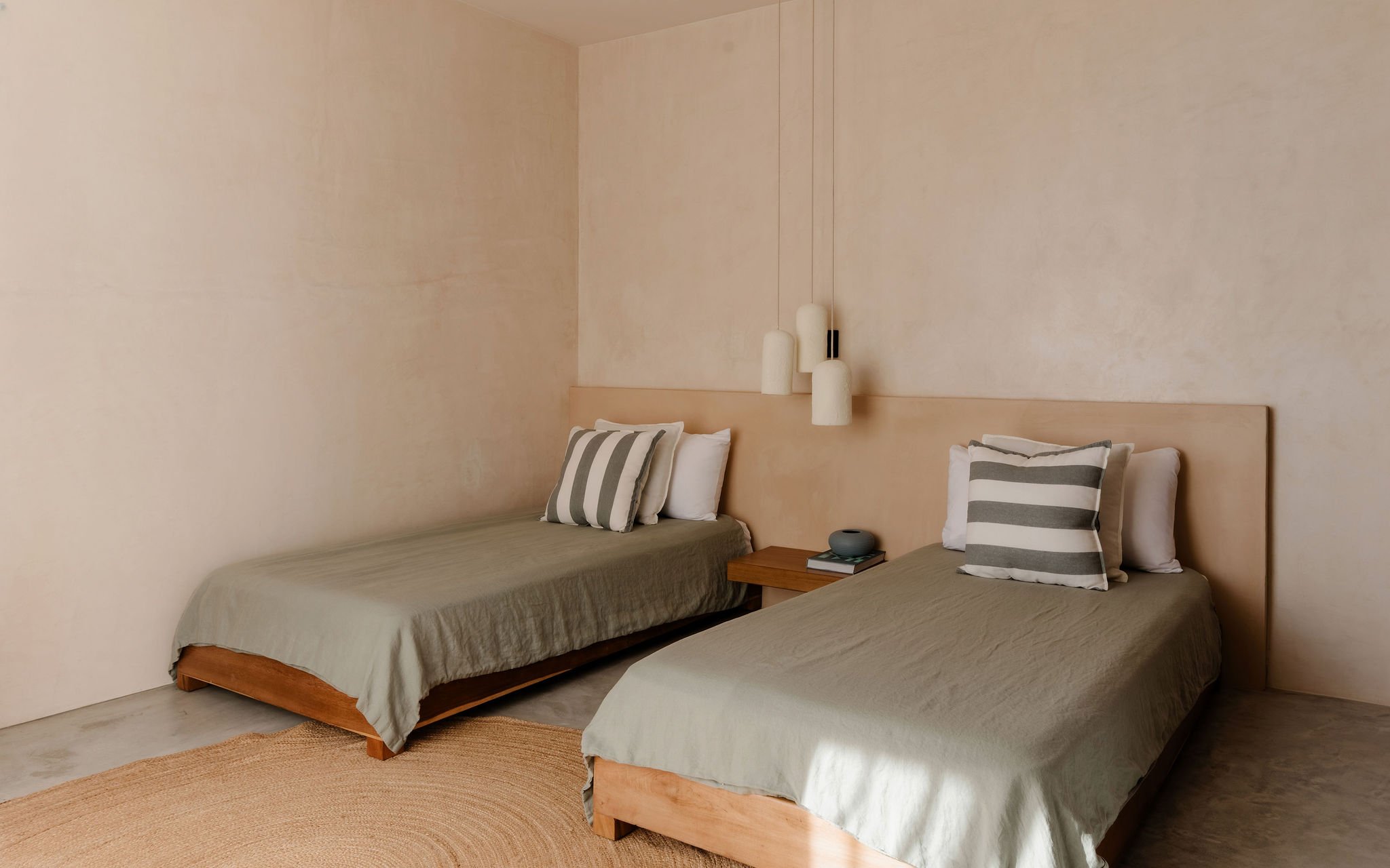 Two twin beds with beige bedspreads and striped pillows, a nightstand with a small blue bowl and a book, and three hanging pendant lights against a pale pink wall in a minimalist bedroom.