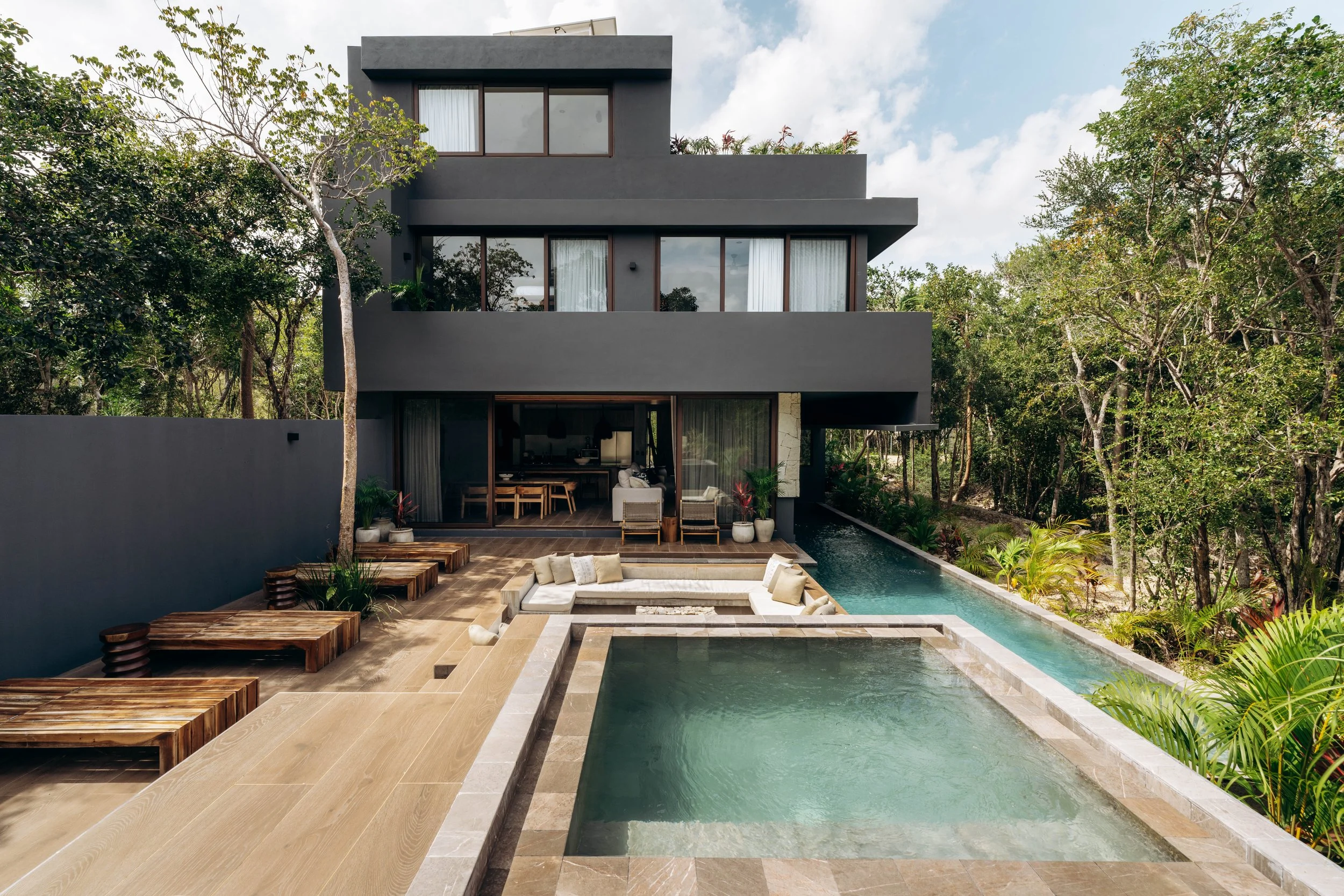 Modern three-story house with large glass windows, outdoor patio area, swimming pool, and lush greenery surrounding the property.