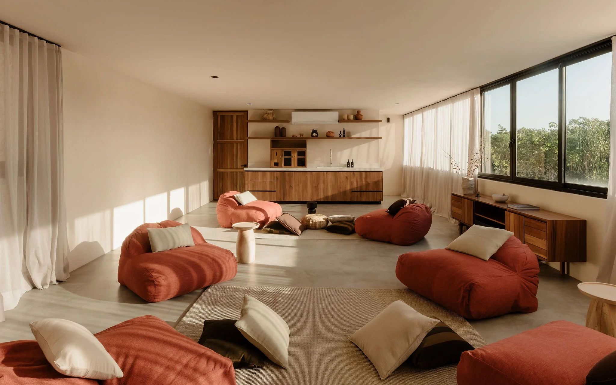 Living room with large windows, beige curtains, and minimalist decor including bean bag chairs, cushions, and wooden furniture.