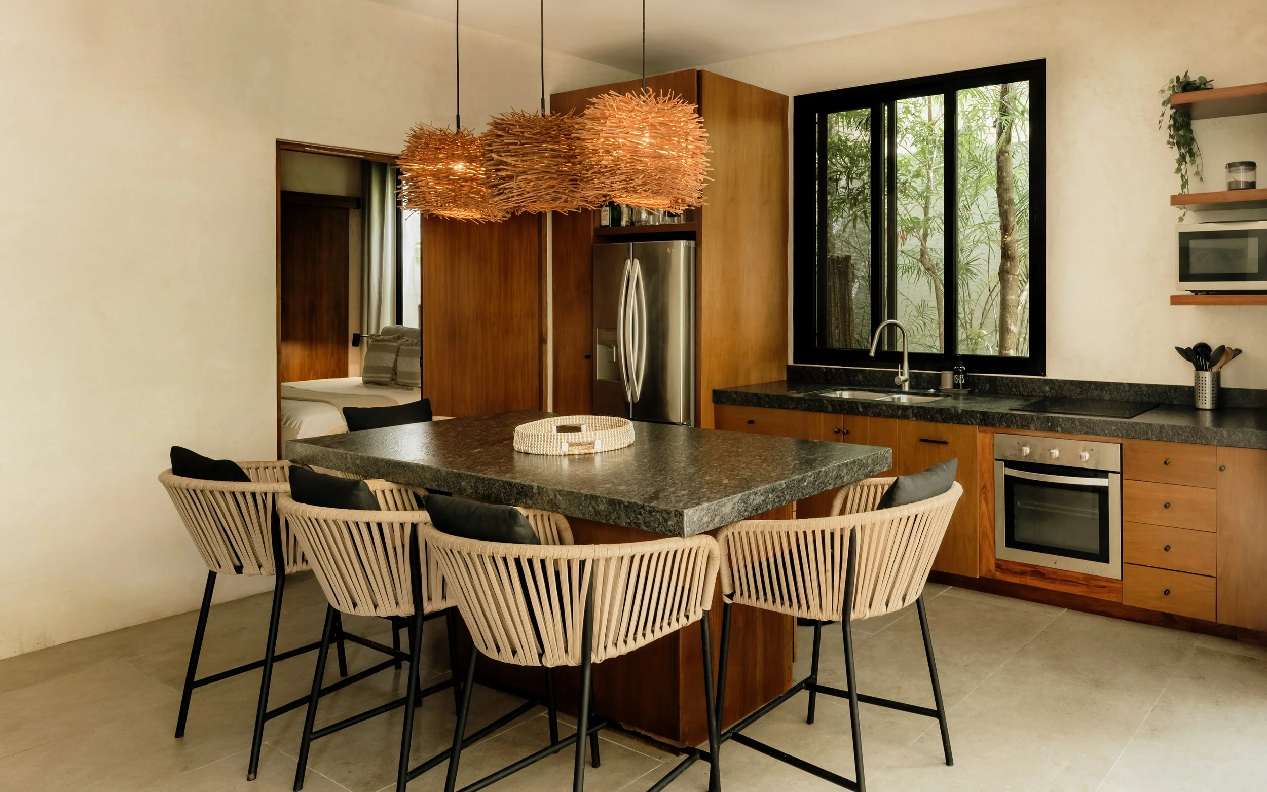 Kitchen with dining area, featuring a dark granite counter, wicker chairs with cushions, wooden cabinets, stainless steel refrigerator, black framed window, and a wooden and woven light fixture hanging from the ceiling.