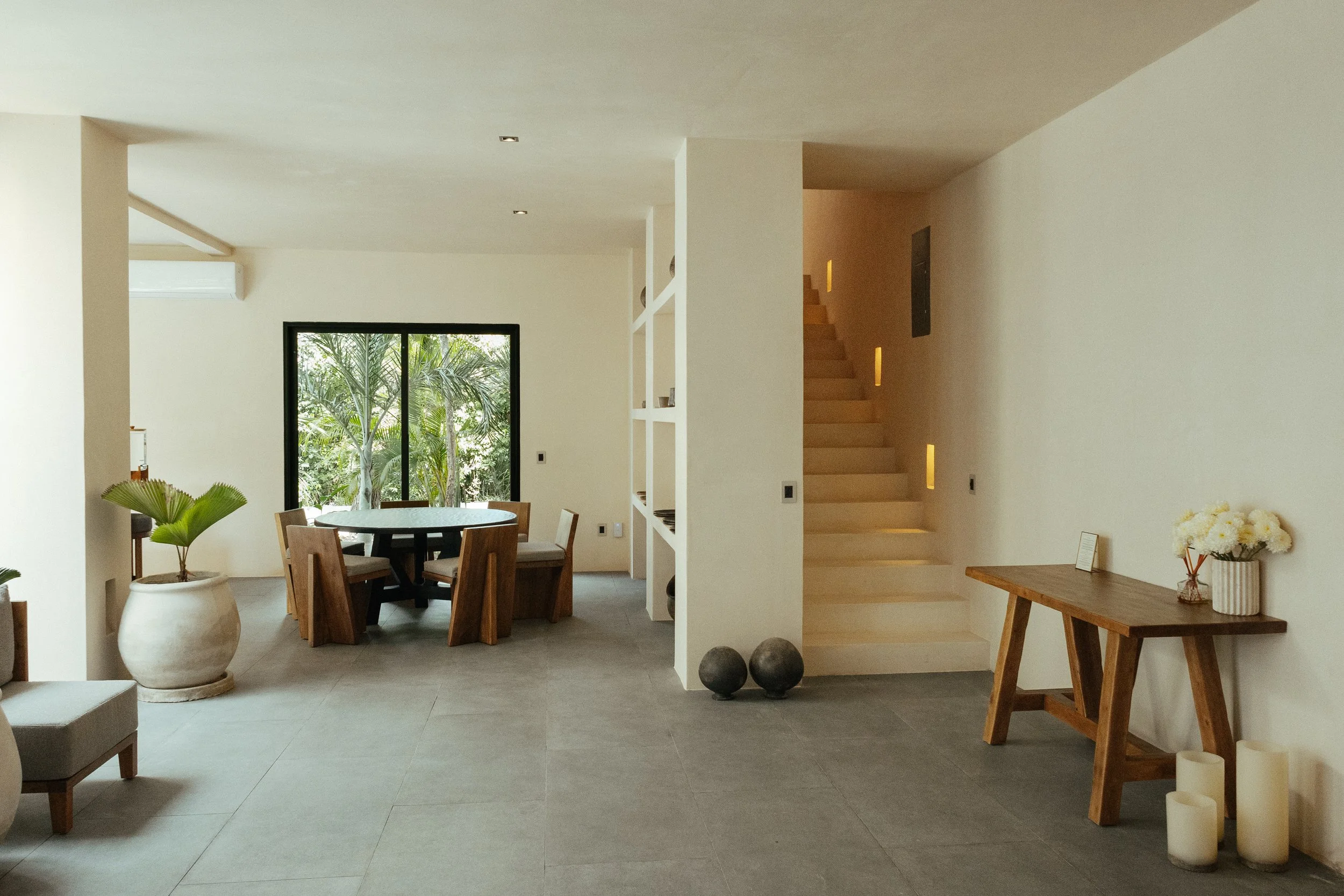 Interior of a modern home with an open plan dining area, white walls, large window showing greenery outside, and minimalistic decor including a wooden table, chairs, potted plants, and candles.