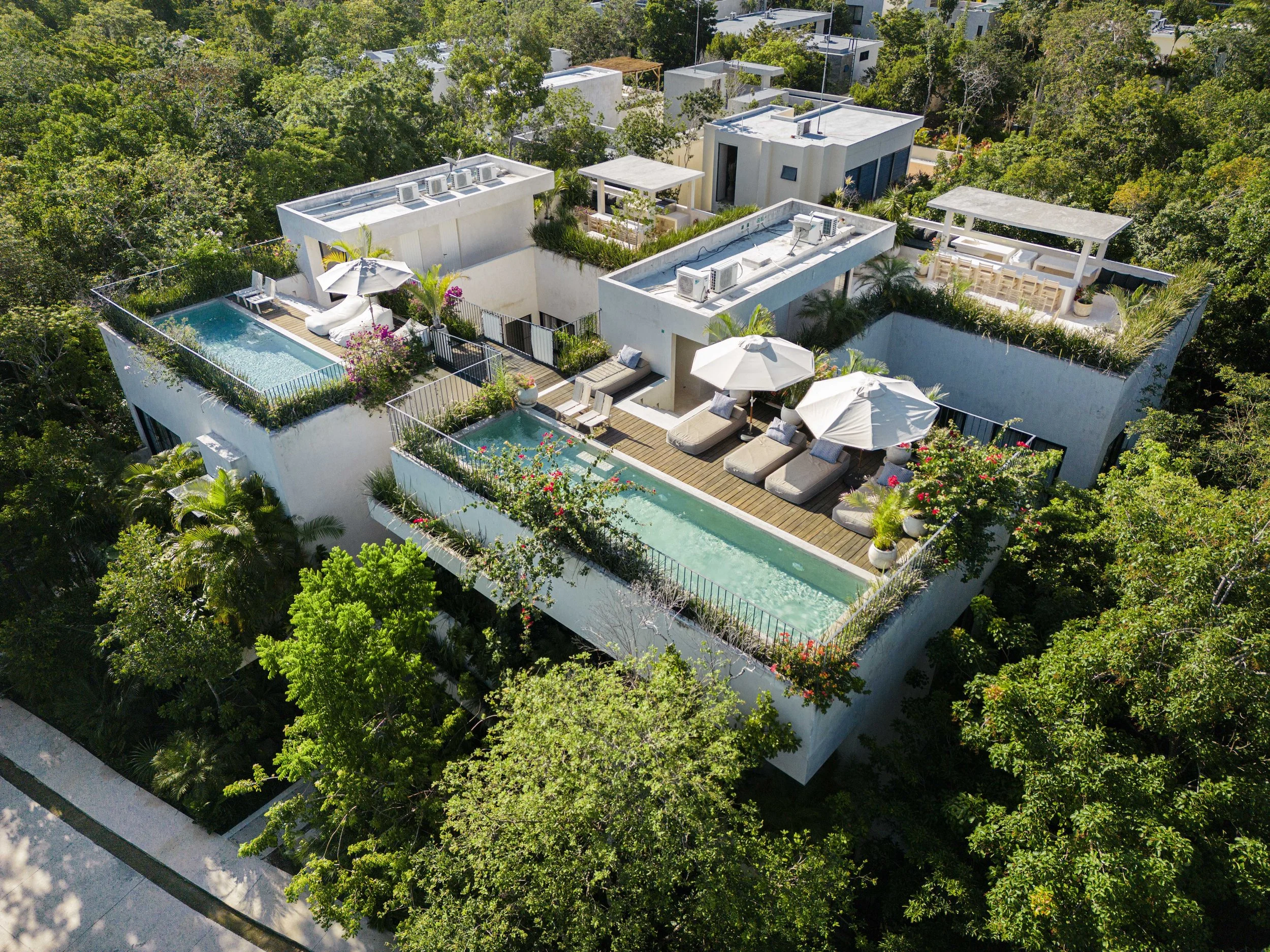 Aerial view of modern rooftop apartments with pools, surrounded by lush green trees.