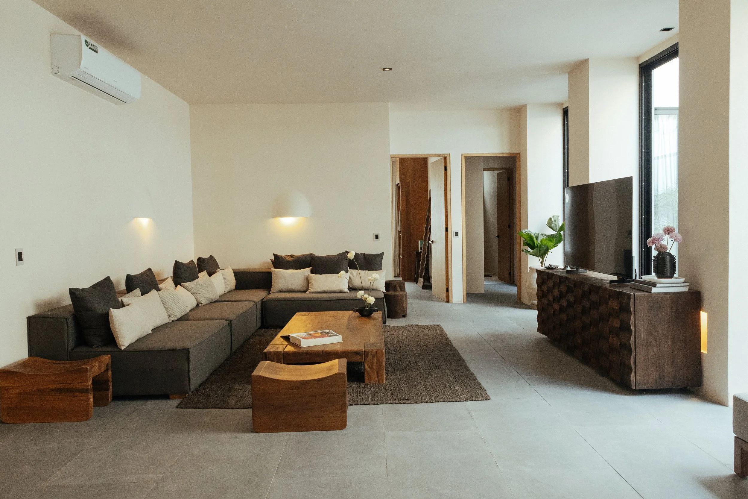 Living room with a gray sectional sofa with white and black pillows, a wooden coffee table with a magazine, a rug, a TV on a wooden stand, potted plants, and large windows letting in natural light.