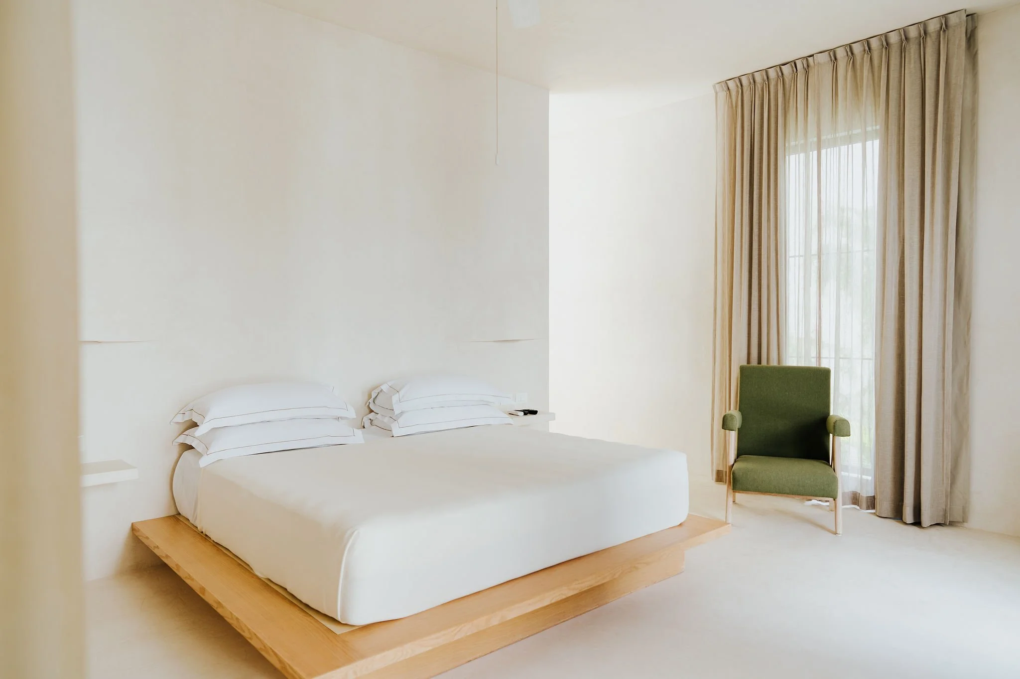 Minimalist hotel room with a low wooden bed frame, white bedding, stacked pillows, and a small side table. There's a green armchair near a large window with sheer curtains, allowing natural light to fill the room.