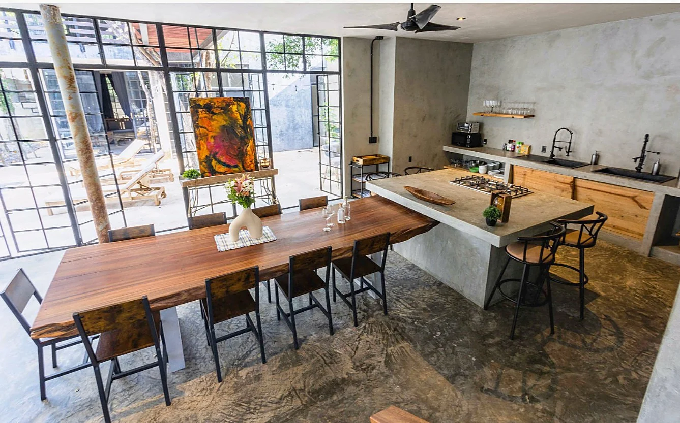 Modern kitchen with a large wooden dining table, black chairs, concrete countertops, black sinks, and an open outdoor area with lounge chairs and outdoor furniture.