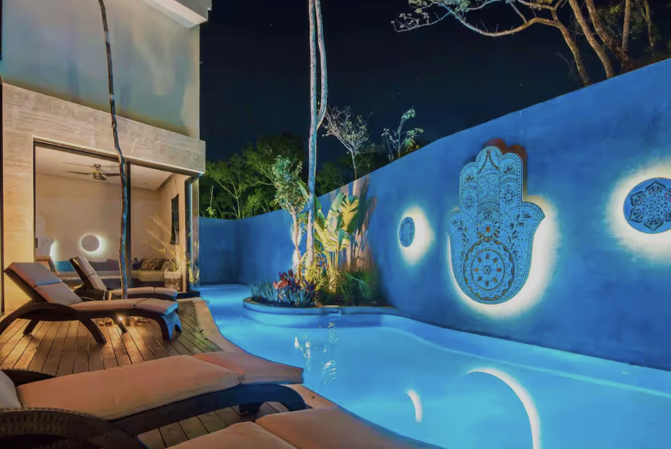 Night view of a backyard pool illuminated with blue lighting, surrounded by lounge chairs, a garden with trees and plants, and a wall with decorative lighting featuring a Hamsa hand design and geometric patterns.
