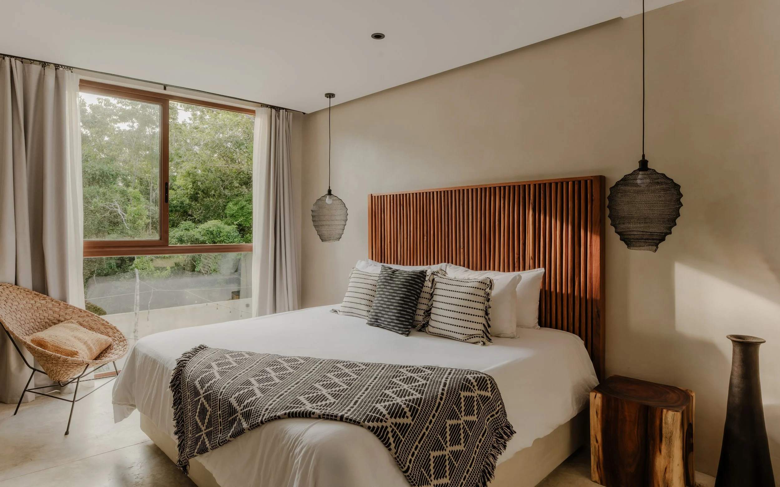 A bedroom with a large window showing greenery outside, a bed with white linens and black-and-white pillows, a wooden headboard with slats, and two hanging pendant lights on either side of the bed.