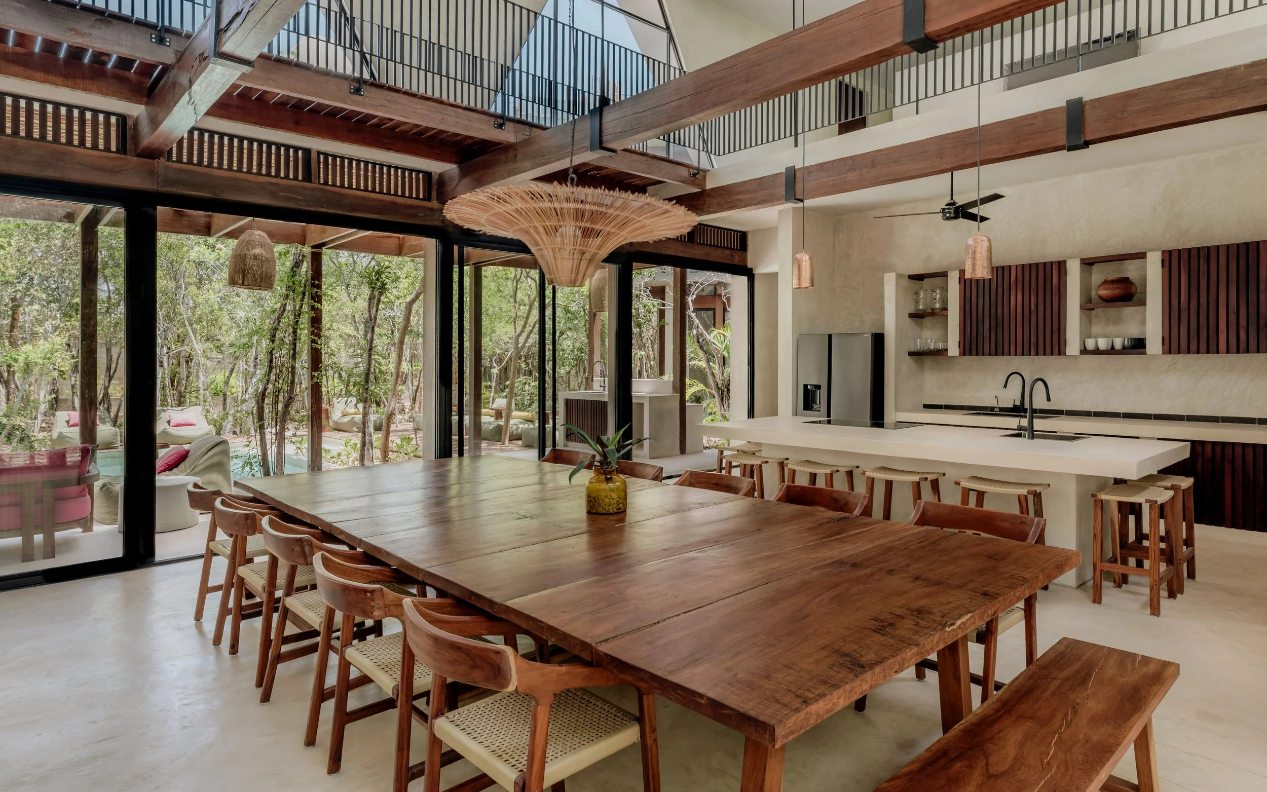 Interior view of a modern, open-concept living space with a dining table, wood accents, and large windows showing an outdoor greenery area.
