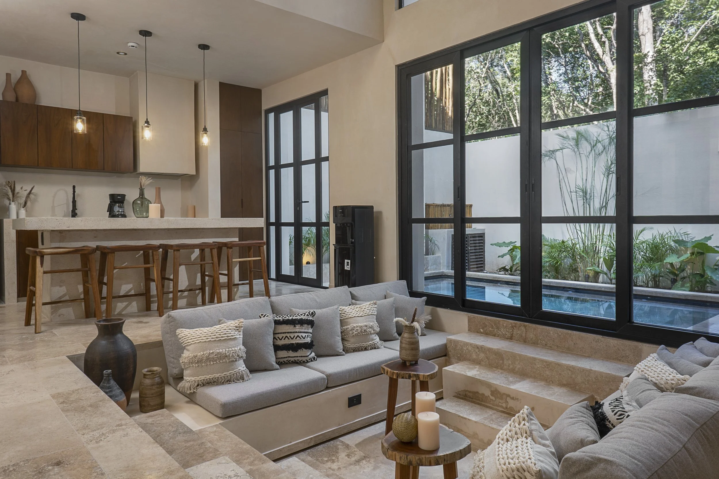 Modern living room with large black-framed windows showing a view of trees and greenery outside. The room features a beige sofa with black and white patterned pillows, a small wooden side table with candles, and decorative vases. The kitchen area has wooden cabinets, a white countertop, and pendant lights hanging from the ceiling.
