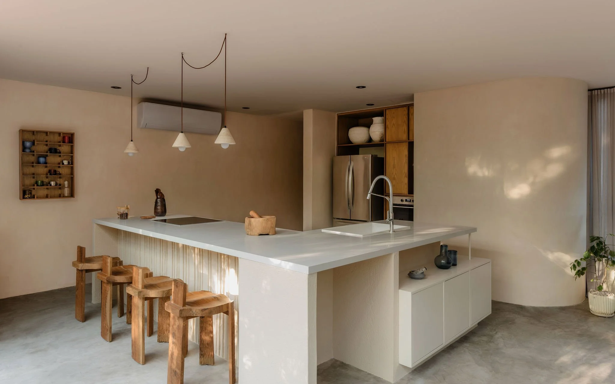 Modern kitchen with white island counter, wooden stools, stainless steel refrigerator, open shelving with pottery, and pendant lights.