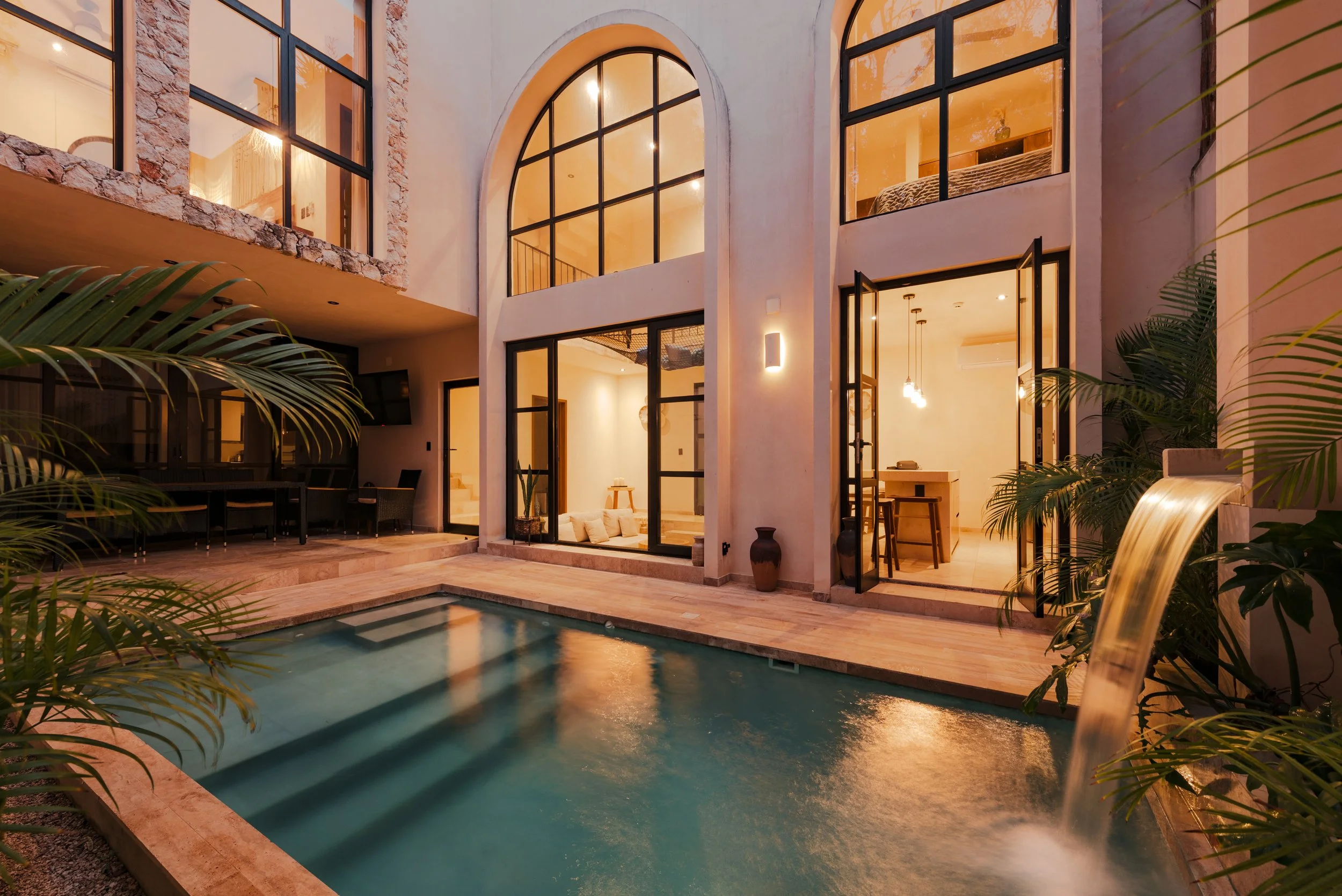 Outdoor view of a modern house with large glass windows and doors, a swimming pool in the foreground, and warm interior lighting, surrounded by indoor plants.