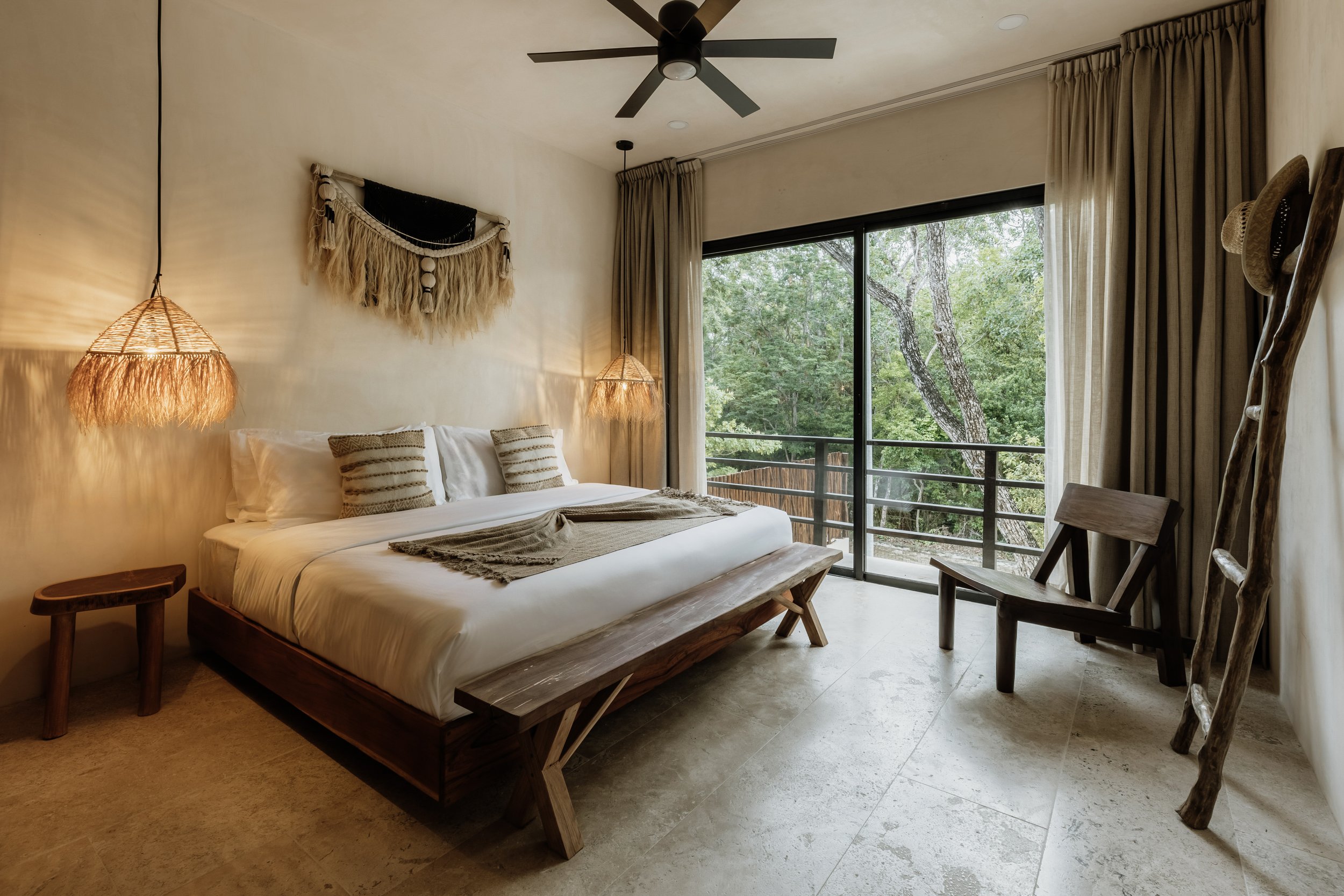 A bedroom with a large window showing green trees outside. The room has a wooden bed with white bedding, decorative pillows, and two hanging bedside lamps with wicker shades. There is a small wooden side table, a wooden chair, a decorative wall hanging, ceiling fan, and beige curtains.