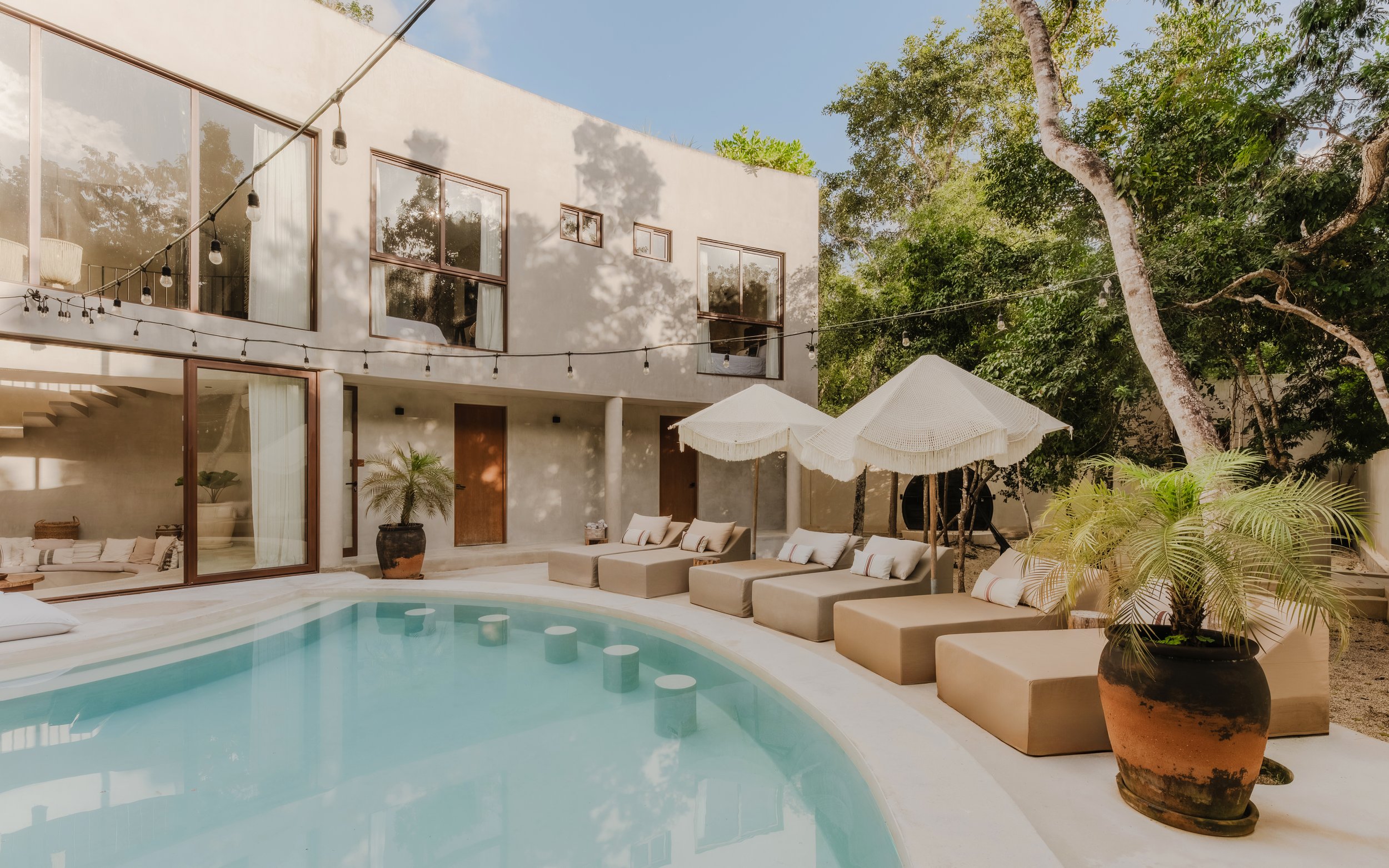 Modern house with large glass windows and patio area, featuring a swimming pool, beige lounge chairs with cushions, large potted plants, and umbrellas, surrounded by tall trees under a clear blue sky.