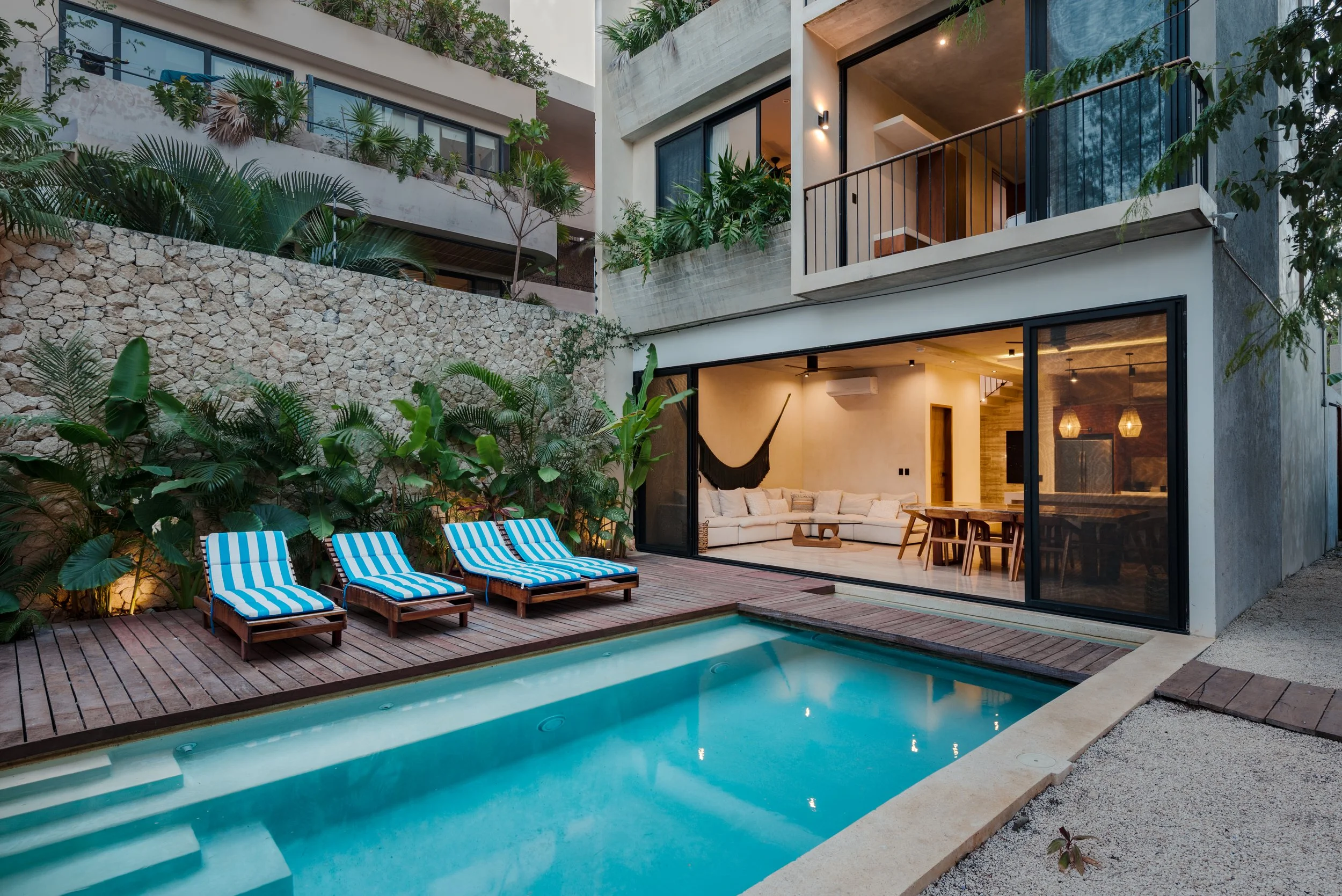 Modern residential building with outdoor pool area, three striped lounge chairs, lush green plants, large windows, and a visible interior living space with a sectional sofa and dining table.